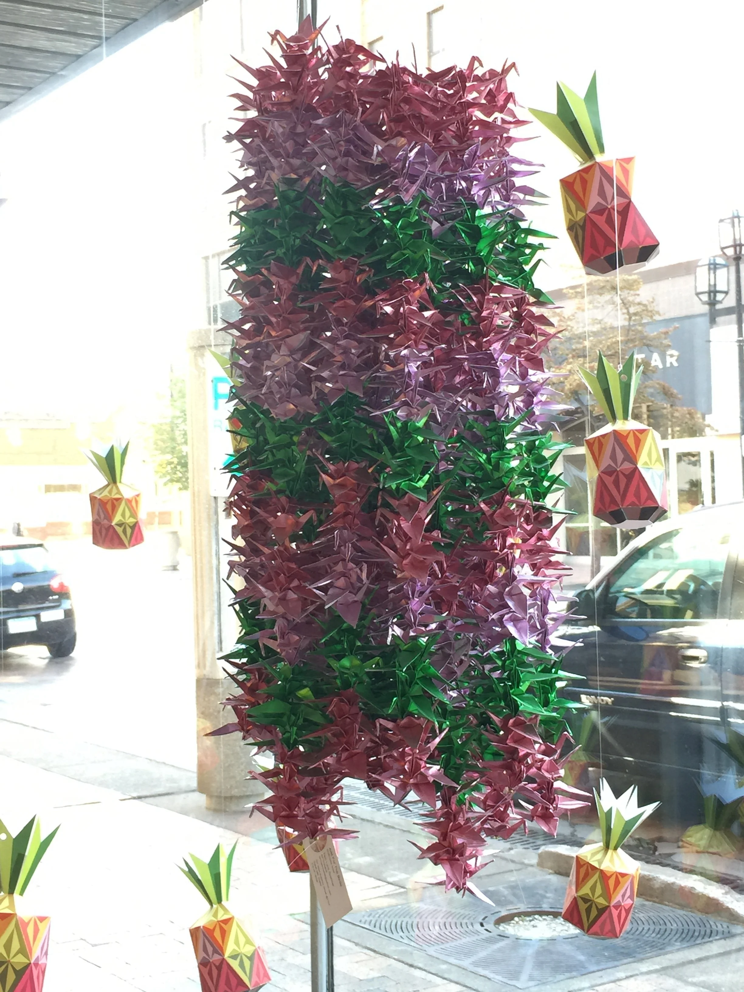 Decorative display with origami pink and purple plant and pineapples on a window, with cars outside.