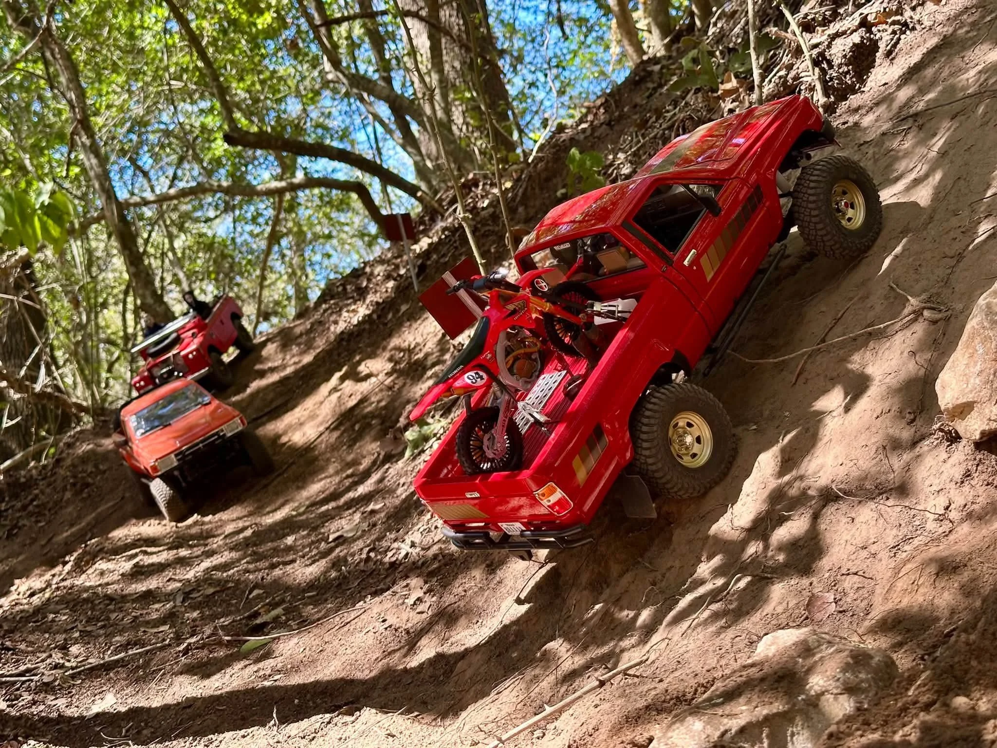 A red RC Crawler with a dirt bike in the truck bed going up an embankment in the woods.
