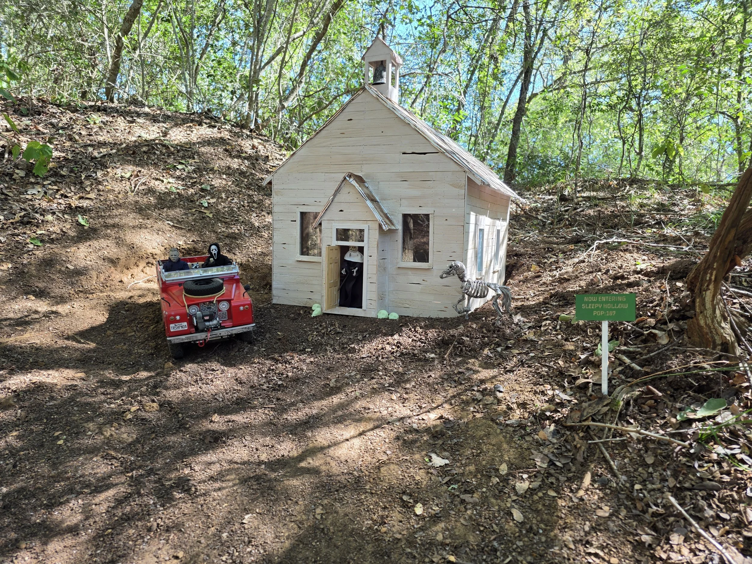 A red scale RC crawler parked beside a miniature haunted schoolhouse on a wooded trail.