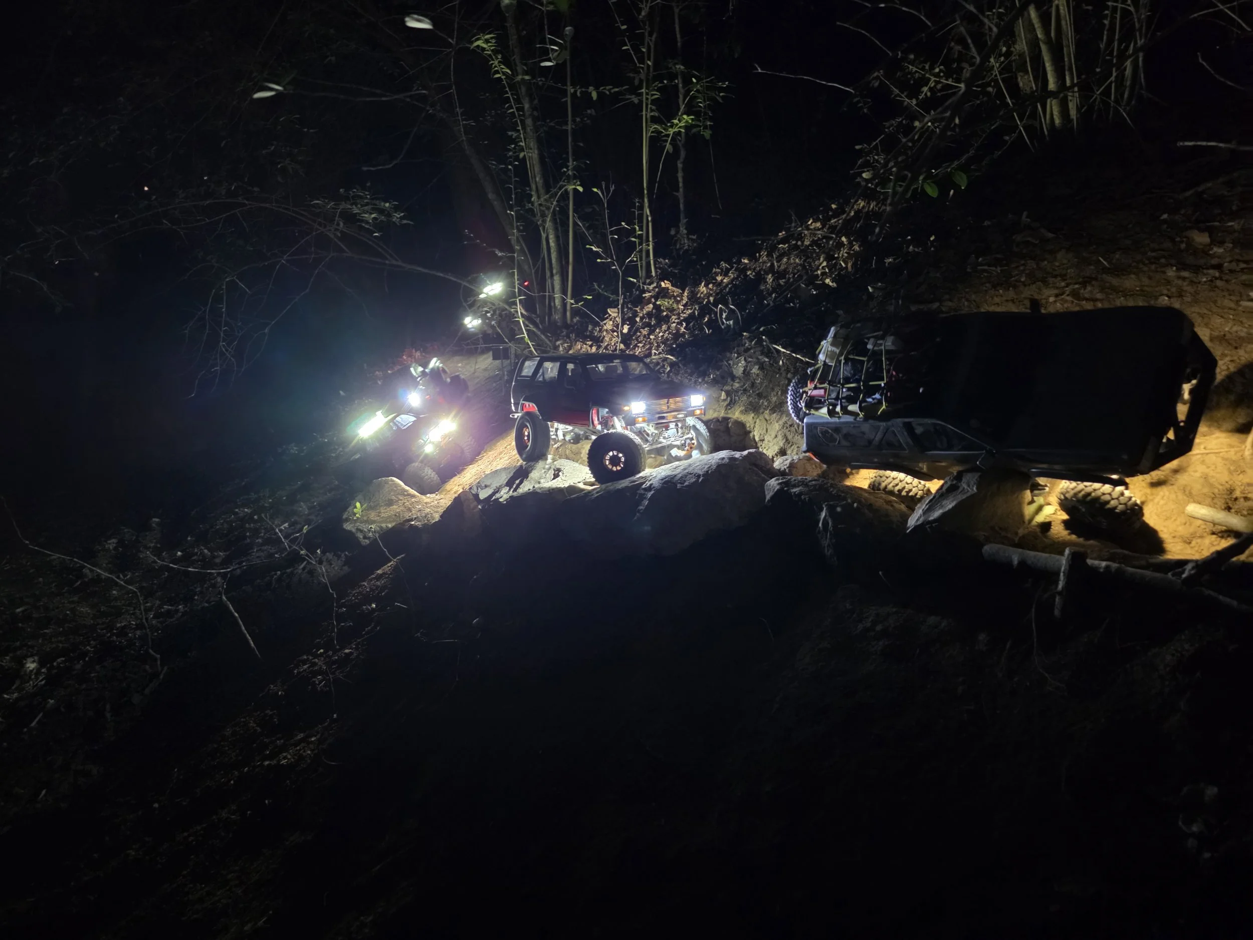 Convoy of Scale RC vehicles driving down a scale dirt road at night with headlights on.