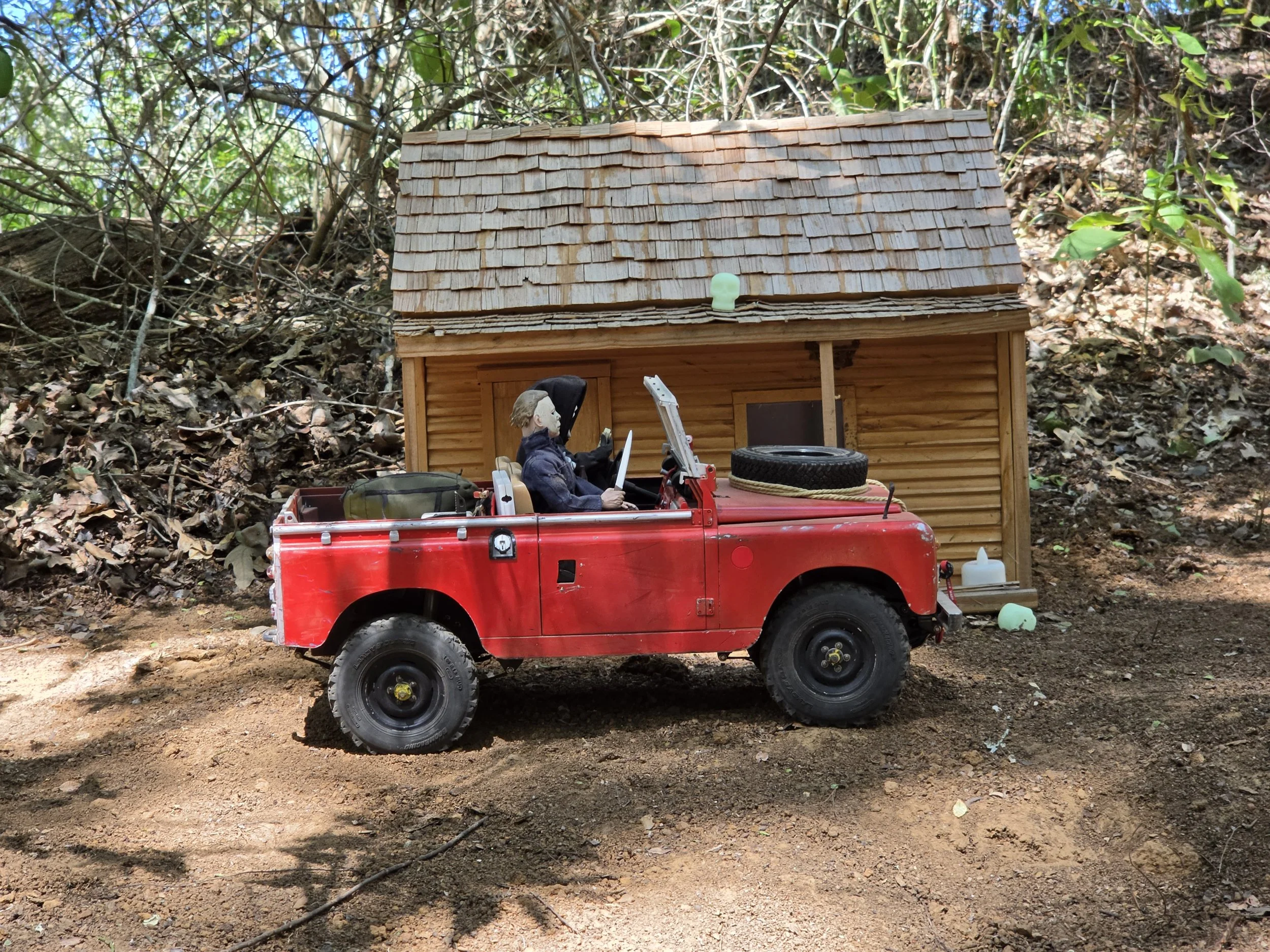 A red RC Crawler Boom truck in front of a miniature building on a dirt road.  The Boom truck hosts Halloween figures in the front seats.