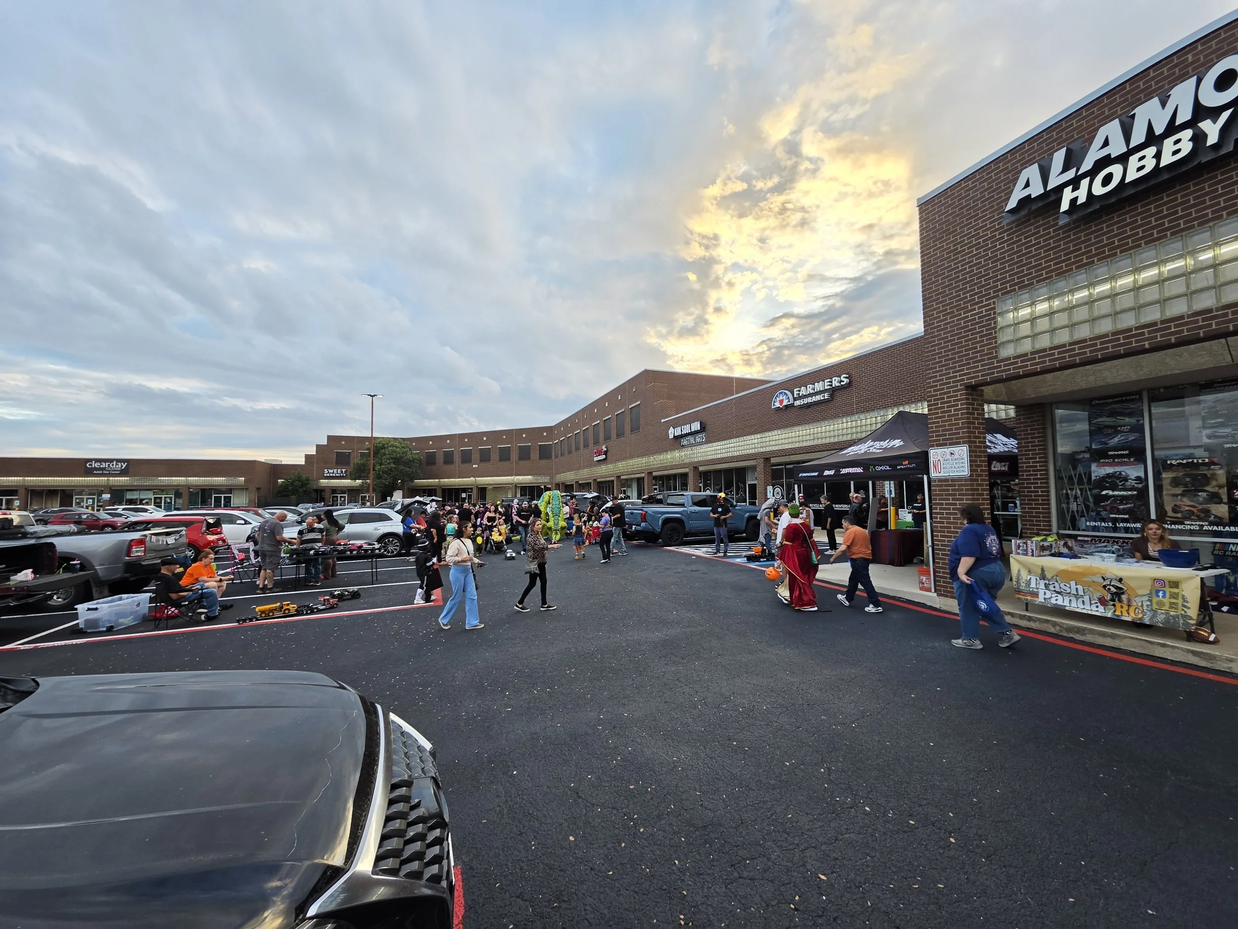 People walking through the parking lof of Alamo City Hobby Shop for the annual Trunk or Treak.  Blue sky with fluffly clouds and sunlight shining through backdrops the lot.