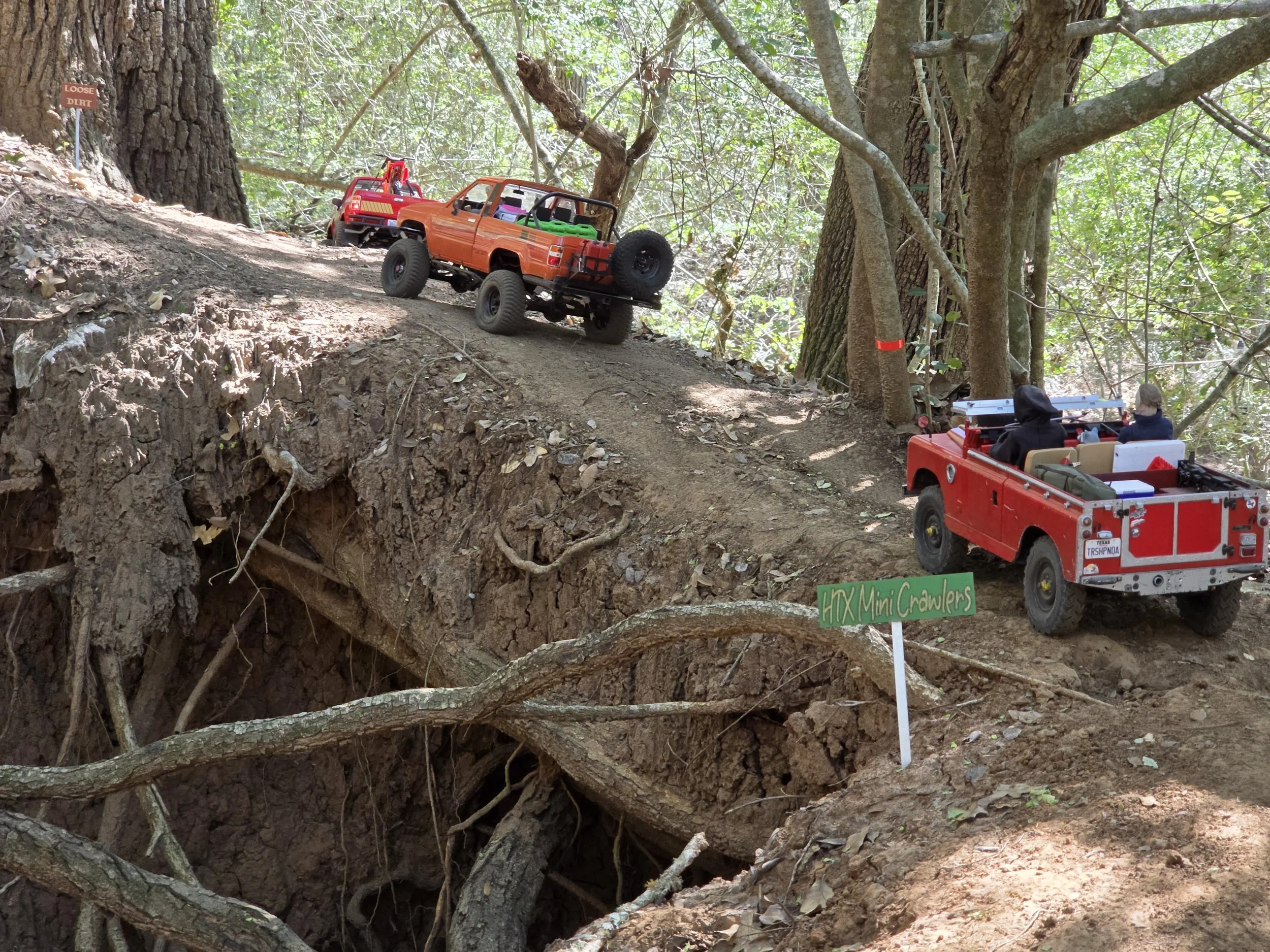 RC crawlers driving up a dirt road with exposed roots.  Miniature HTX Mini Crawler sign in the foreground with trees and greenery in the background.