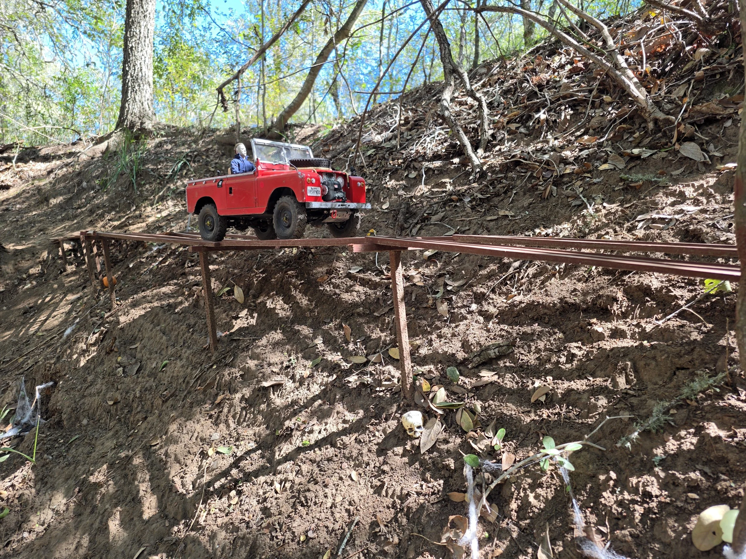 A red Boom RC truck riding along a miniature wooden bridge surrounded by wooded area.