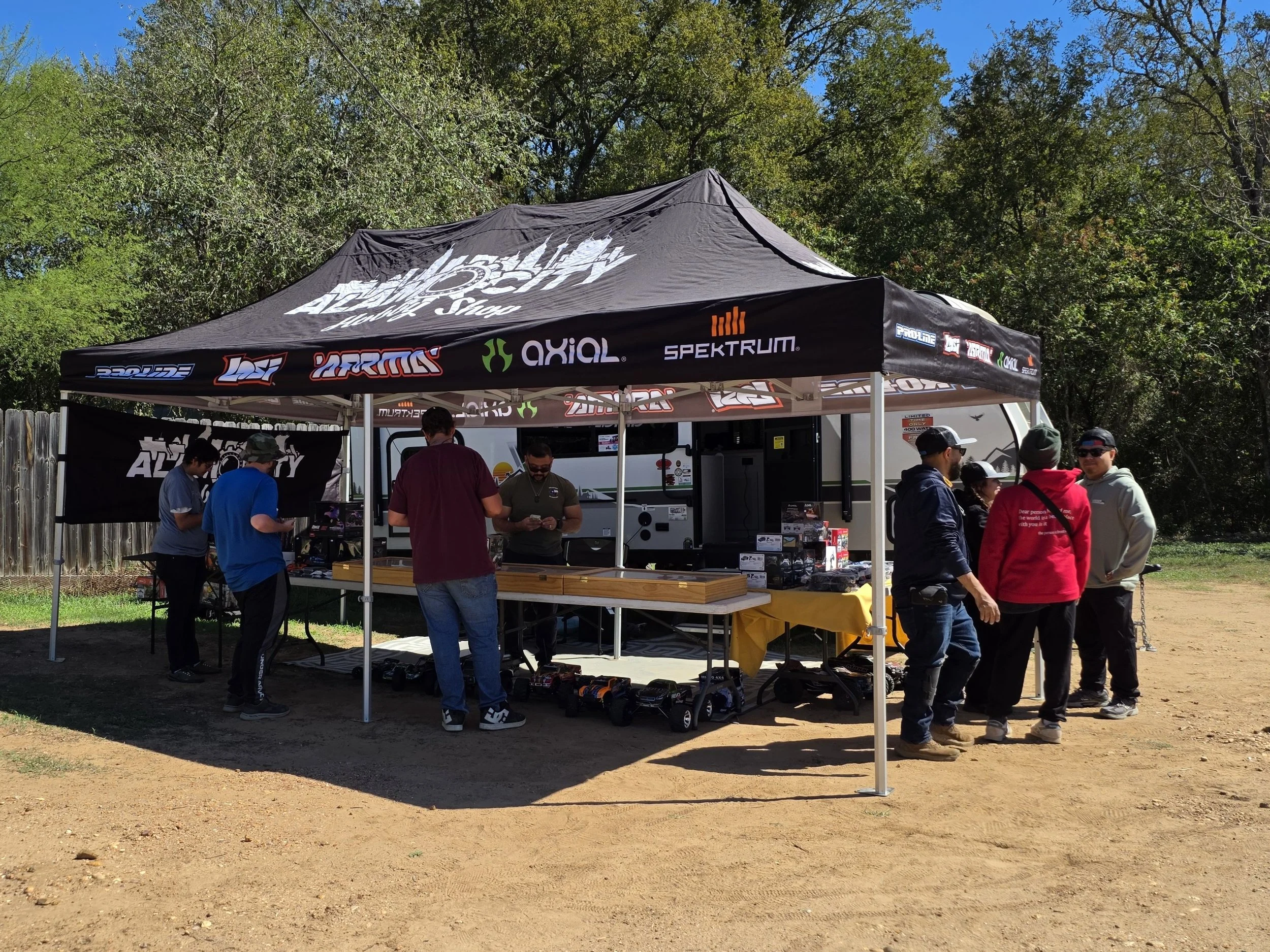 The Alamo City Hobby Shop canopy featuring the San Antonio skyline.  The canopy sits on a dirt road with green trees in the background.  Visitors and shoppers stand at the booth.