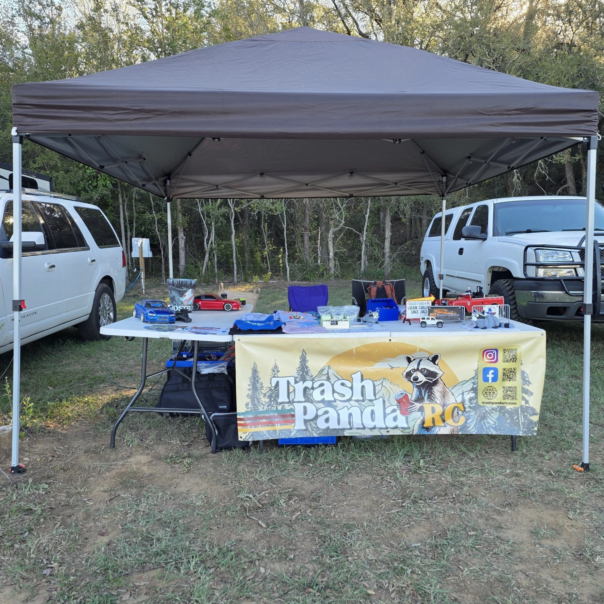 The Trash Panda RC outdoor vendor booth featuring a Raccoon banner, RC crawler models, branded merchandise, and accessories beneath a canopy