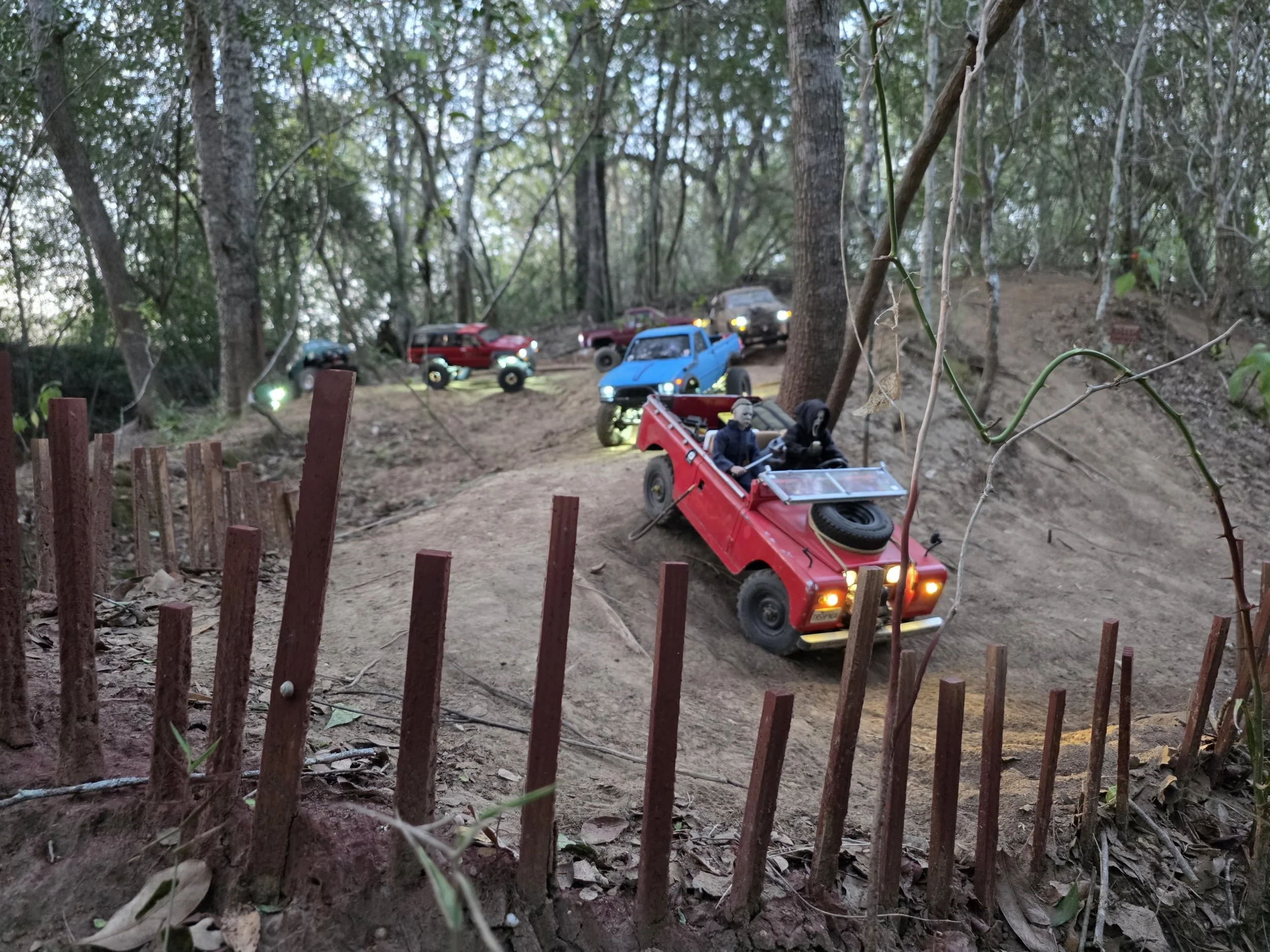Ghost Face driving a red RC Crawler with Michael Myers sitting the passenger seat.  Several other RC vehicles follow on a dirt scale trail.