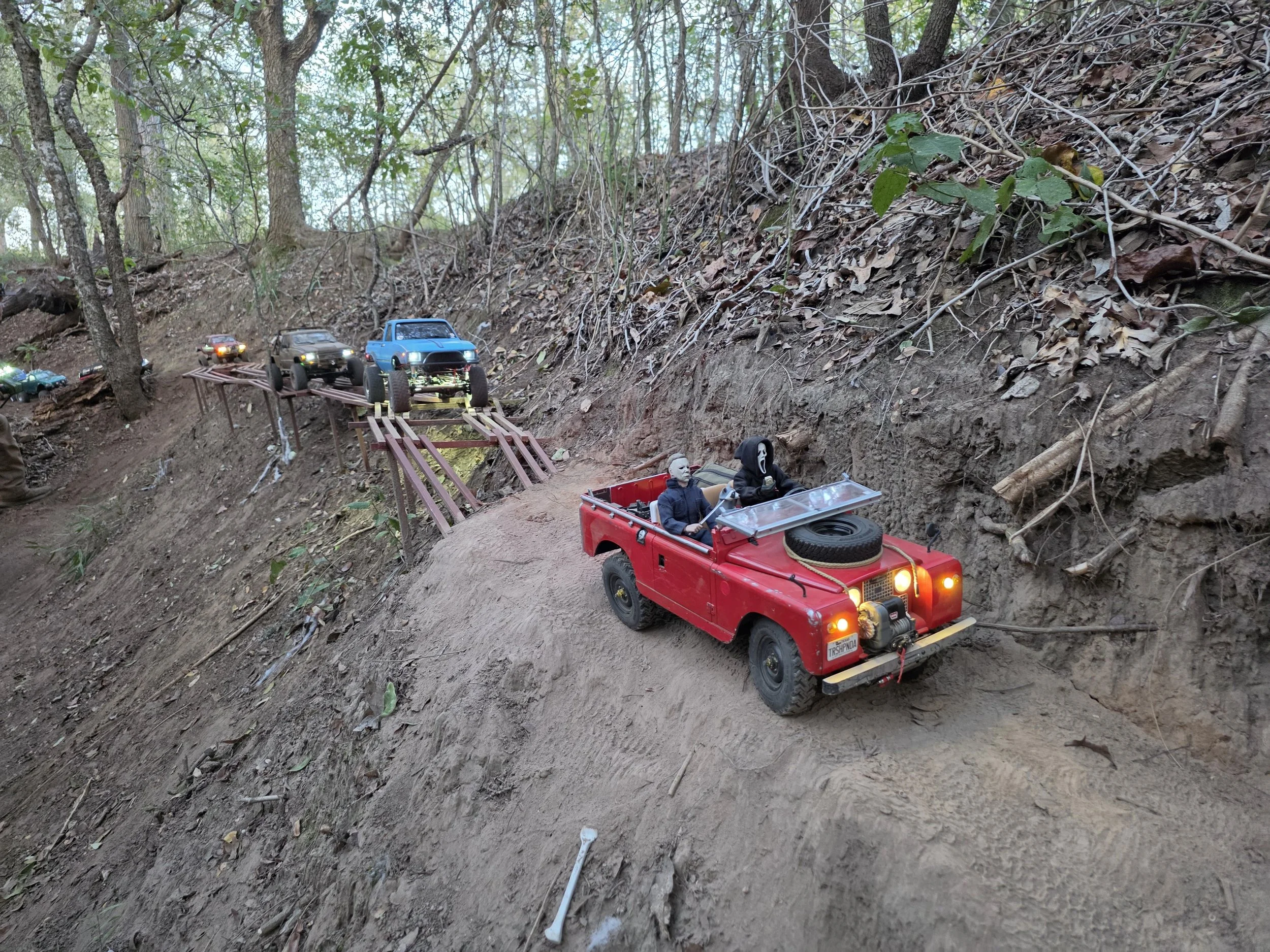 A convoy of RC Scale trucks going over a scale wooden bridge at dusk.  The trucks have their lights on and the lead truck is being driven by Ghost Face with Michael Myers in the drivers seat.
