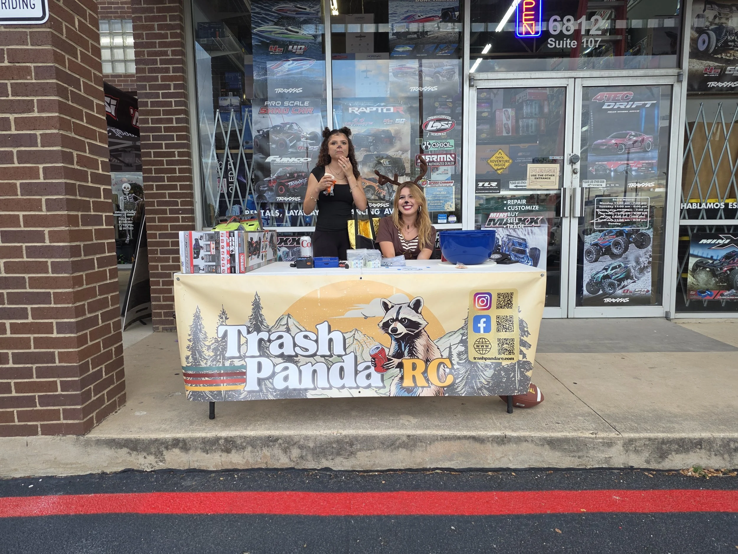 Two girls  in costumes set up at  a table outside the Alamo City Hobby Shop.  The Trash Panda RC logo is on a banner on the table.  One of the girls is dressed as a mouse, the other is a deer.