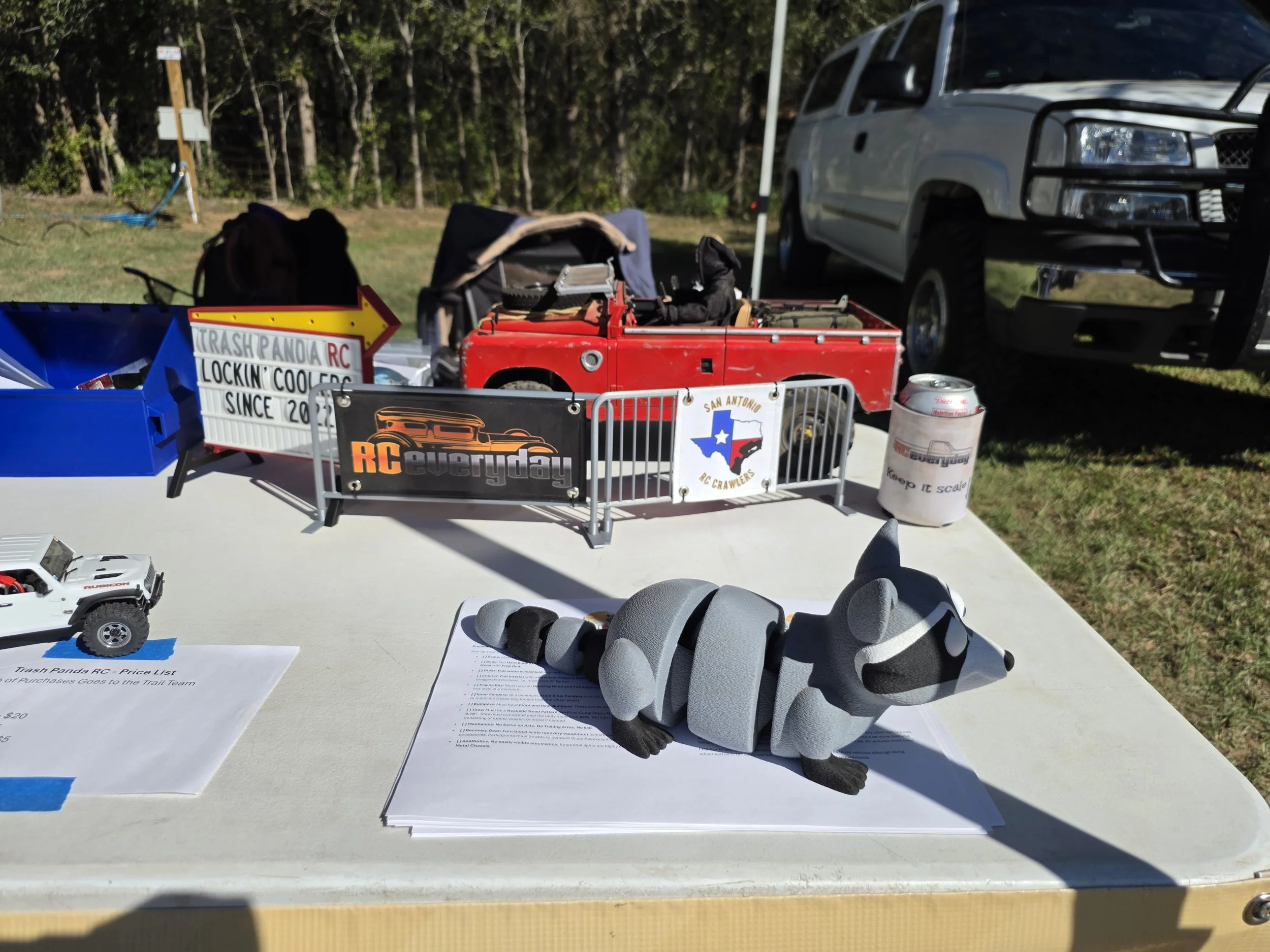 An articulating raccoon sitting on a table in front of a scale barricade with RC everyday and  San Antonio RC Crawler banners.  A red RC car sits behind the barricade.