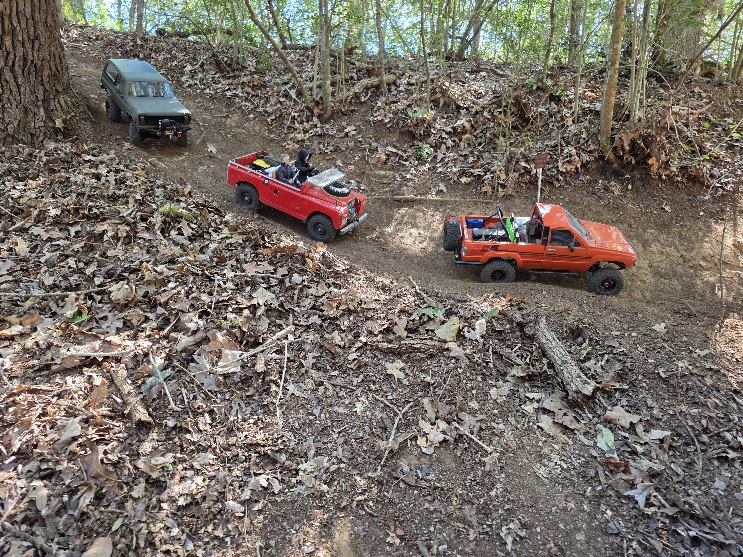 Three RC Crawlers driving along a scale dirt road in a wooded area.
