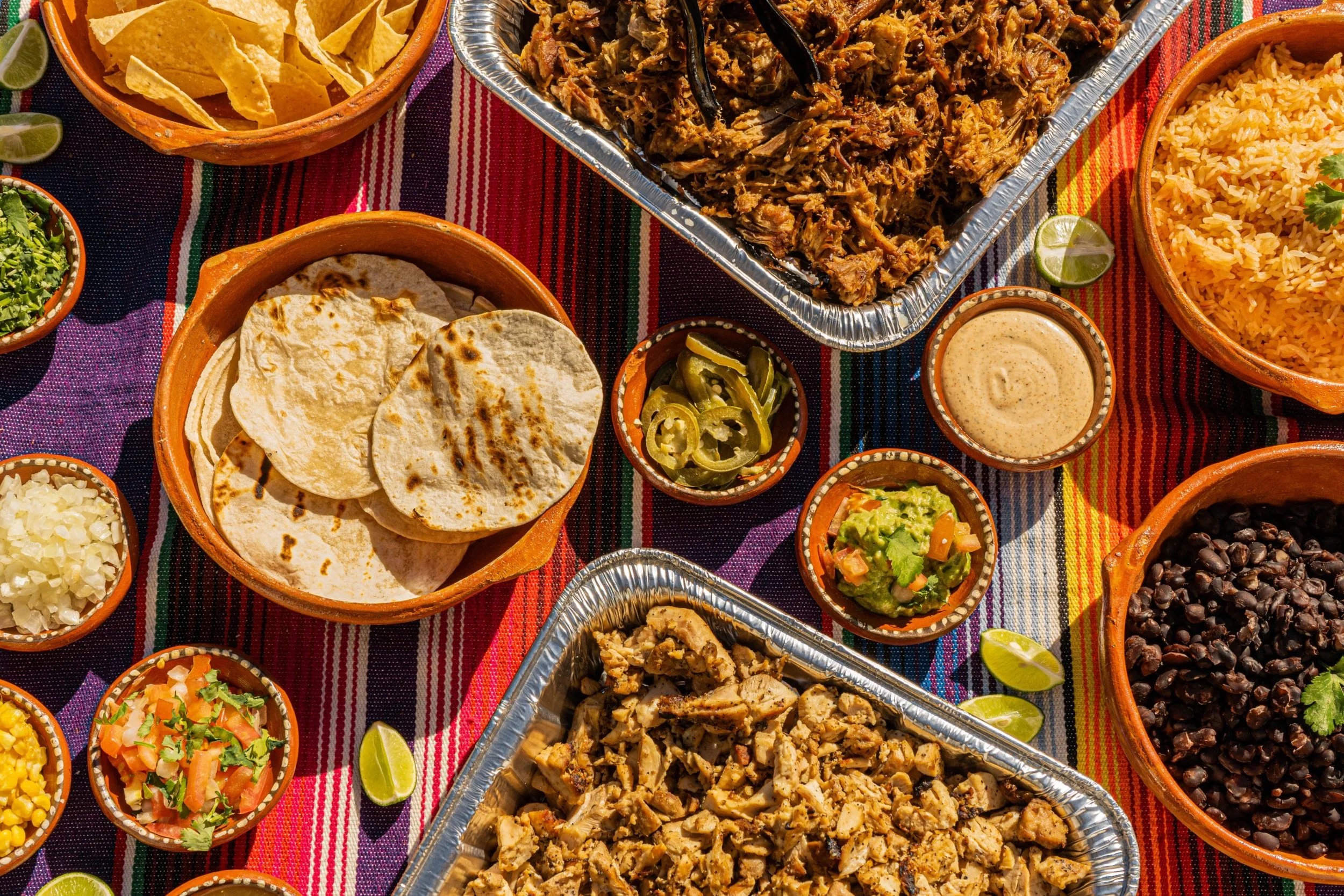 A colorful Mexican feast with dishes including shredded meat, rice, refried beans, guacamole, salsa, tortillas, and lime wedges on a vibrant woven tablecloth.
