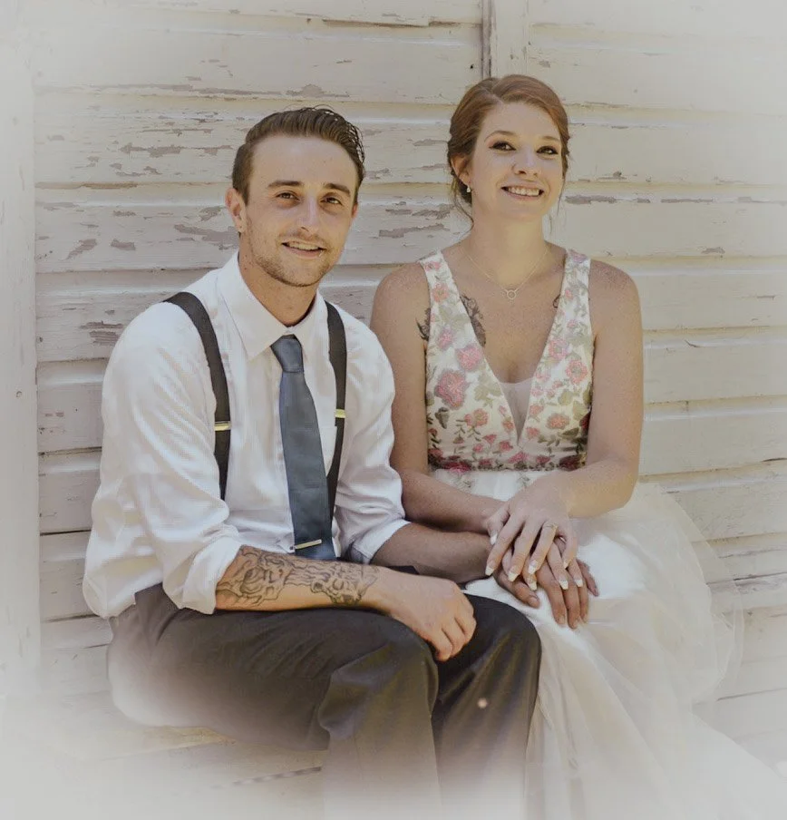 A smiling man and woman sitting close together against a white wooden wall. The man wears a white shirt, tie, suspenders, and has tattoos on his arm. The woman wears a floral top and a white skirt, with her hair up and hands clasped on her lap.