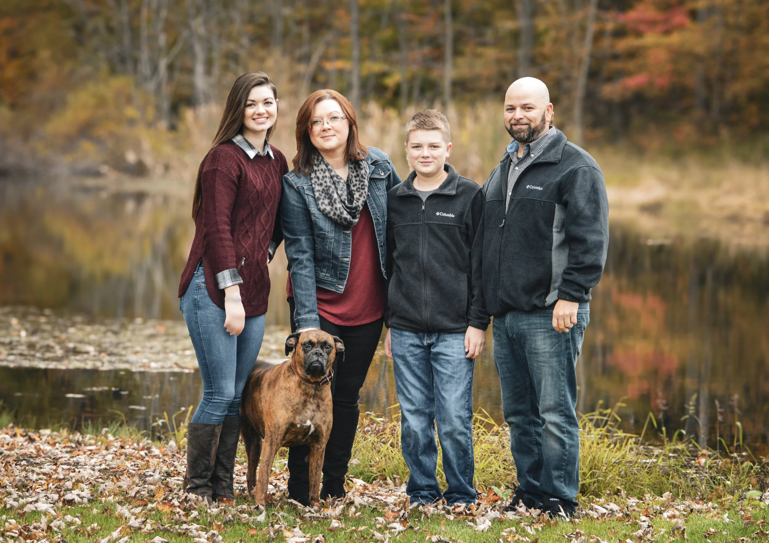A family of four with a dog standing outdoors in an autumn setting near a lake, with colorful fall trees in the background.
