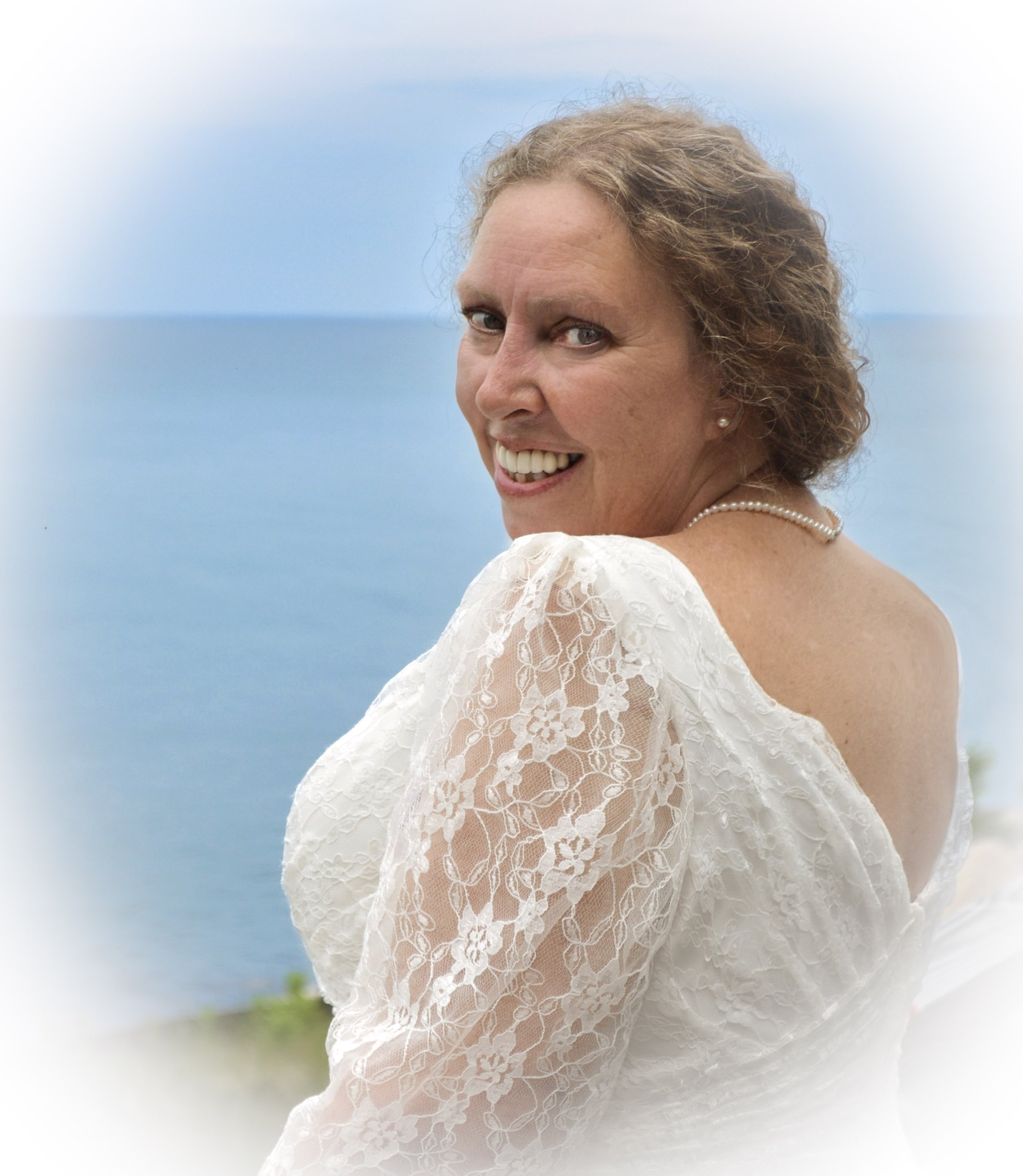Close-up of a smiling woman with curly hair wearing a lace white dress and pearl necklace, against a blue sky background.