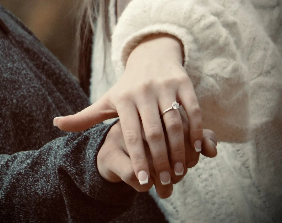 Close-up of two people's hands clasped together, with a woman showing her engagement ring with a large central stone.