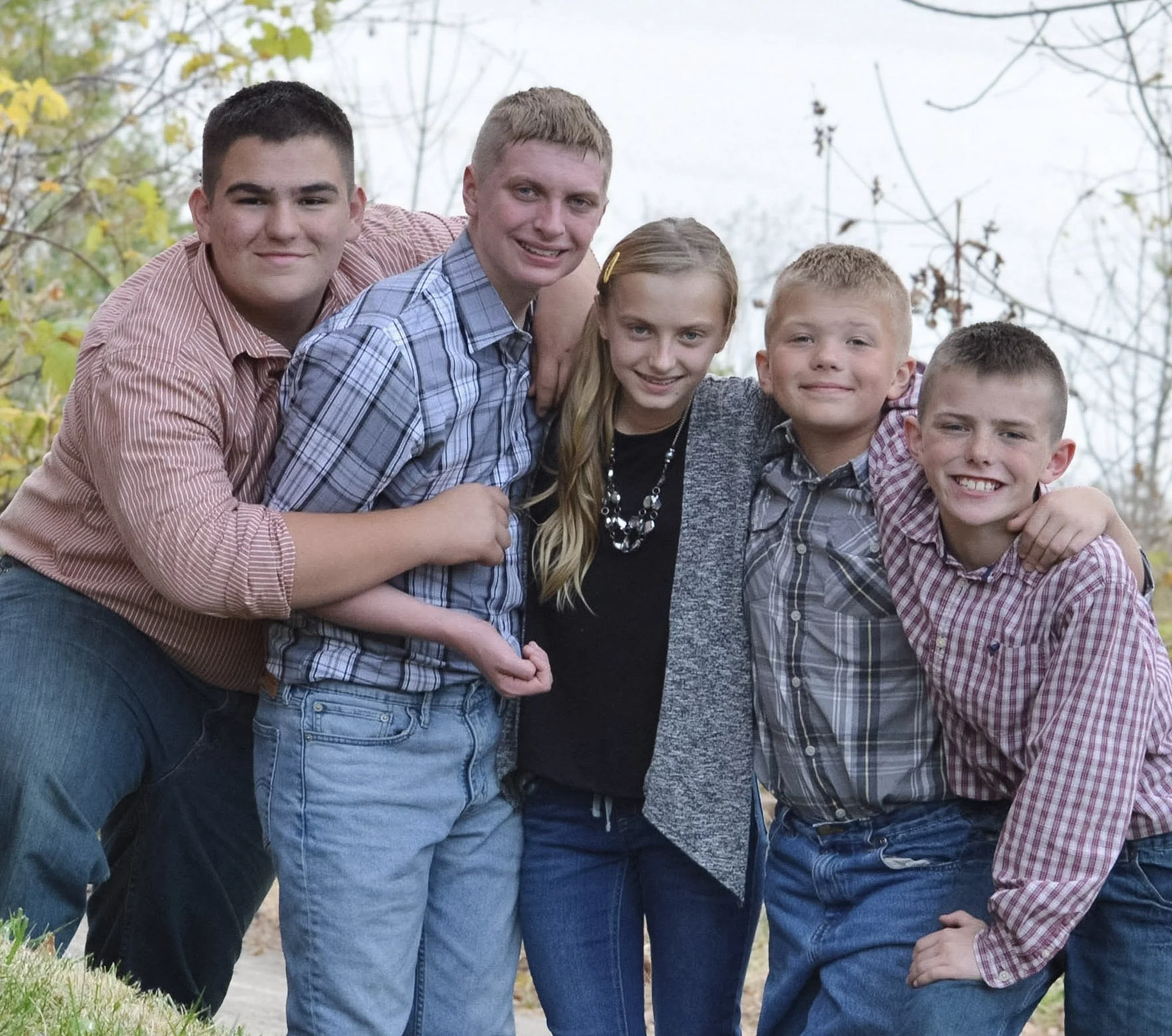 A group of five children, three boys and two girls, smiling and leaning together outdoors during fall, with trees and fallen leaves in the background.