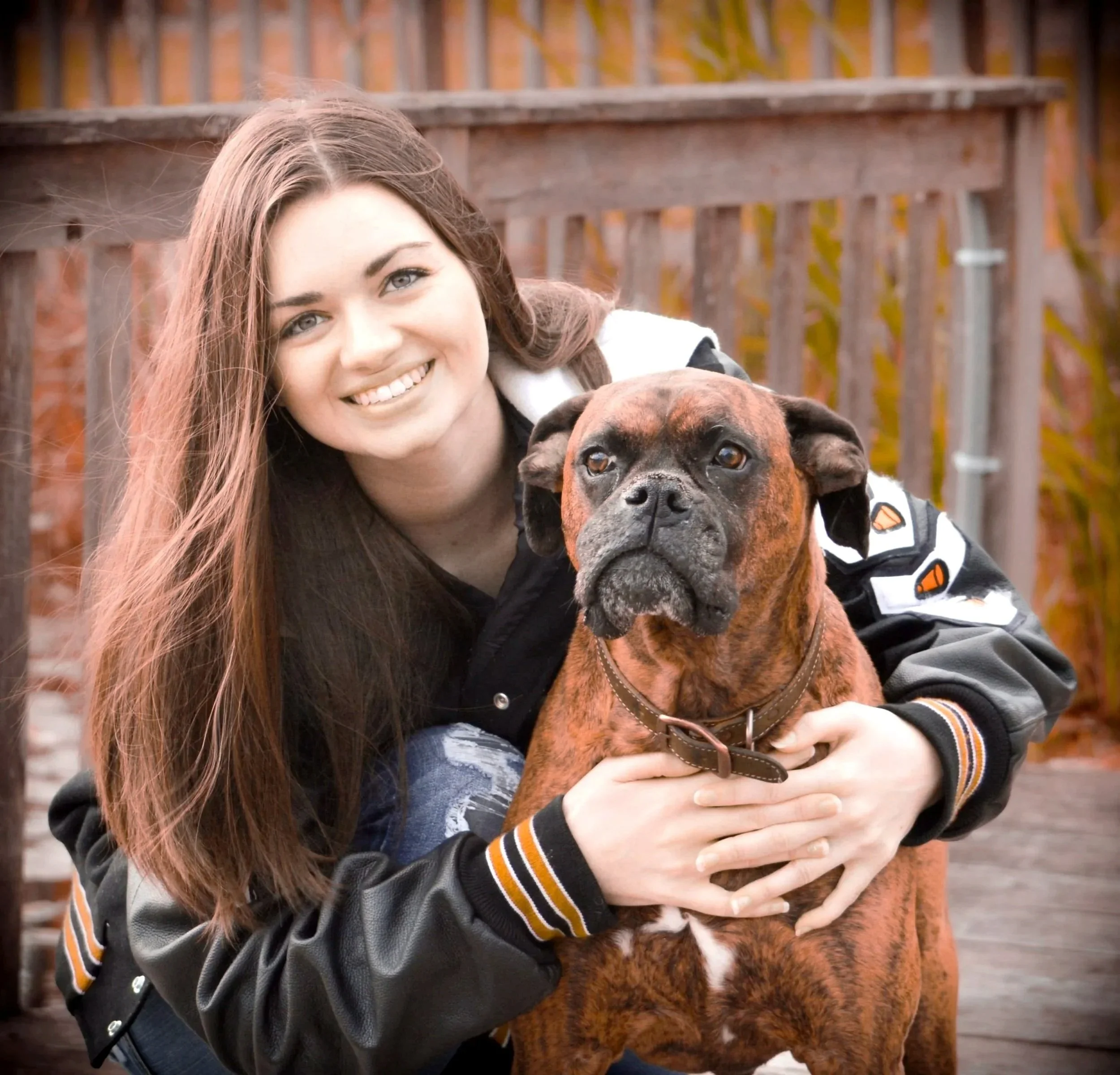A young woman with long brown hair smiling, hugging a brindle-coated dog outdoors, with a wooden fence in the background.