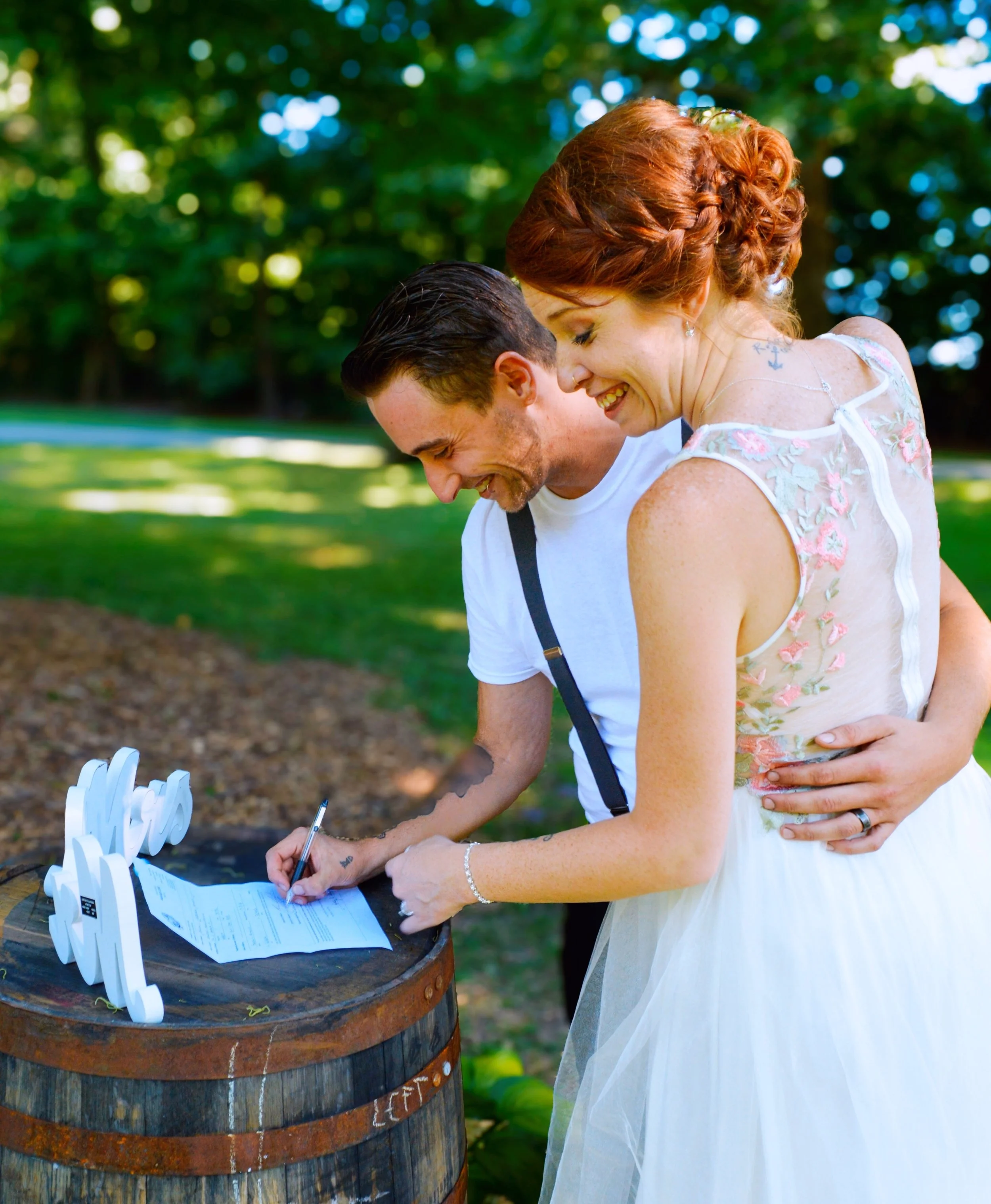 A woman in a white dress with floral details is signing a document on a table outside, with a man next to her. They are smiling and appear happy, with a background of green trees and dappled sunlight.