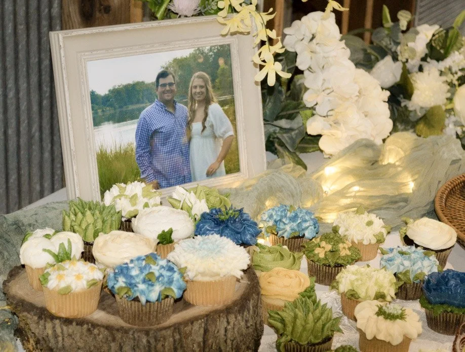 A memorial or tribute display featuring a framed photo of a smiling couple outdoors by a lake, surrounded by numerous decorated cupcakes with floral and nature-themed frosting in white, blue, green, and beige colors, along with white flowers and greenery on a table.