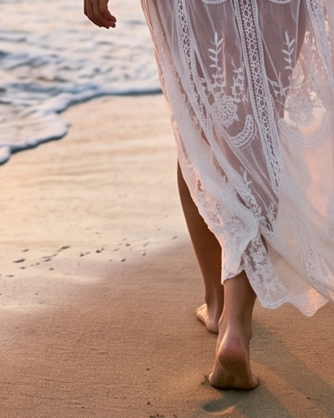 Close-up of barefoot woman walking on the beach at sunset, wearing a white lace dress.