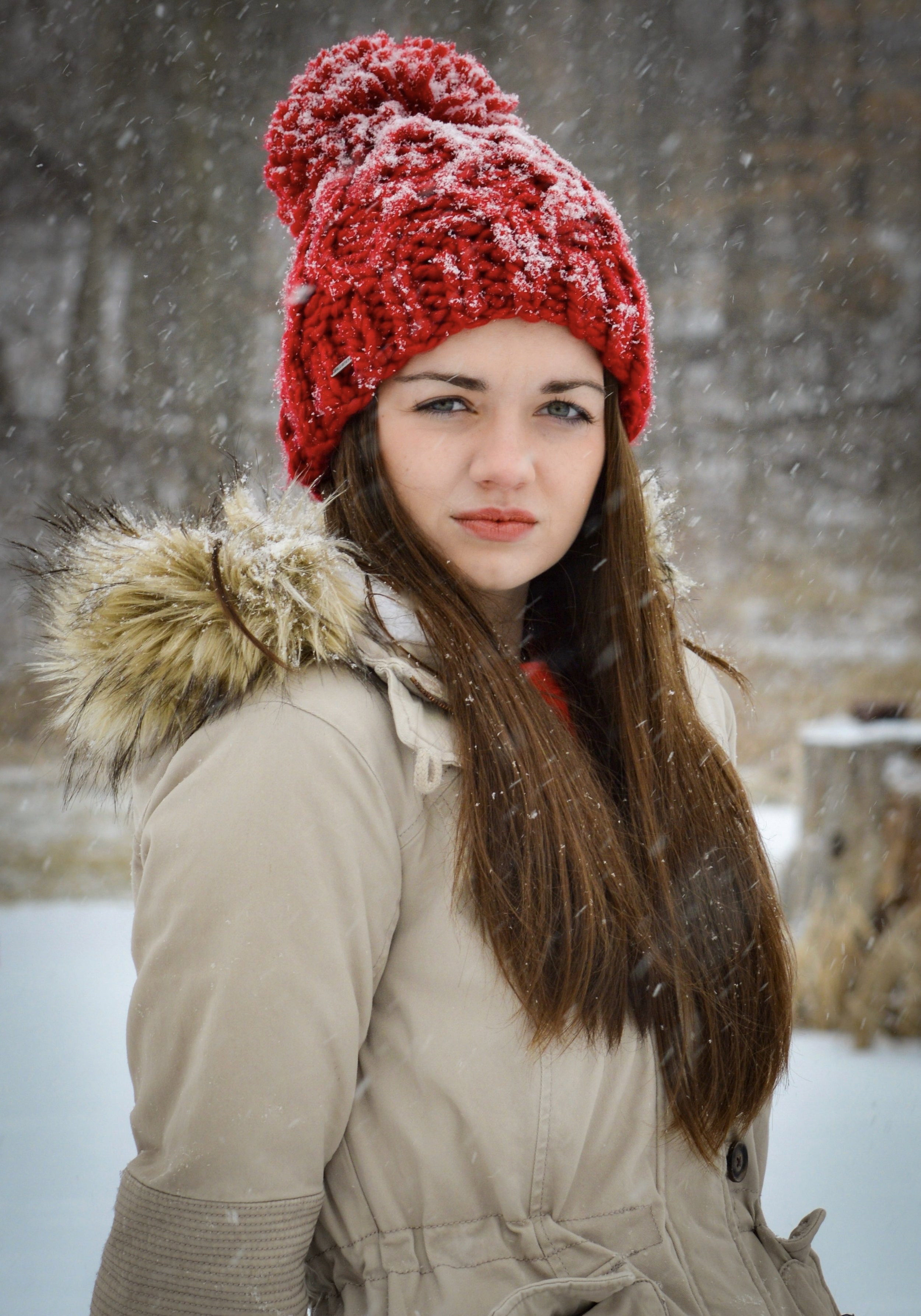 A young woman with brown hair and gray eyes wearing a red knit beanie with snow on it, a white winter coat with a fur-lined hood, outdoors during snowfall.