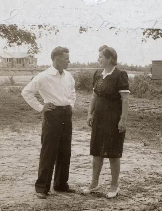 A black-and-white photo of a young man and woman standing outdoors, facing each other, with the man in a white shirt and dark trousers, and the woman in a dark dress and heels. They are on a dirt ground with a rural background, including trees and buildings.