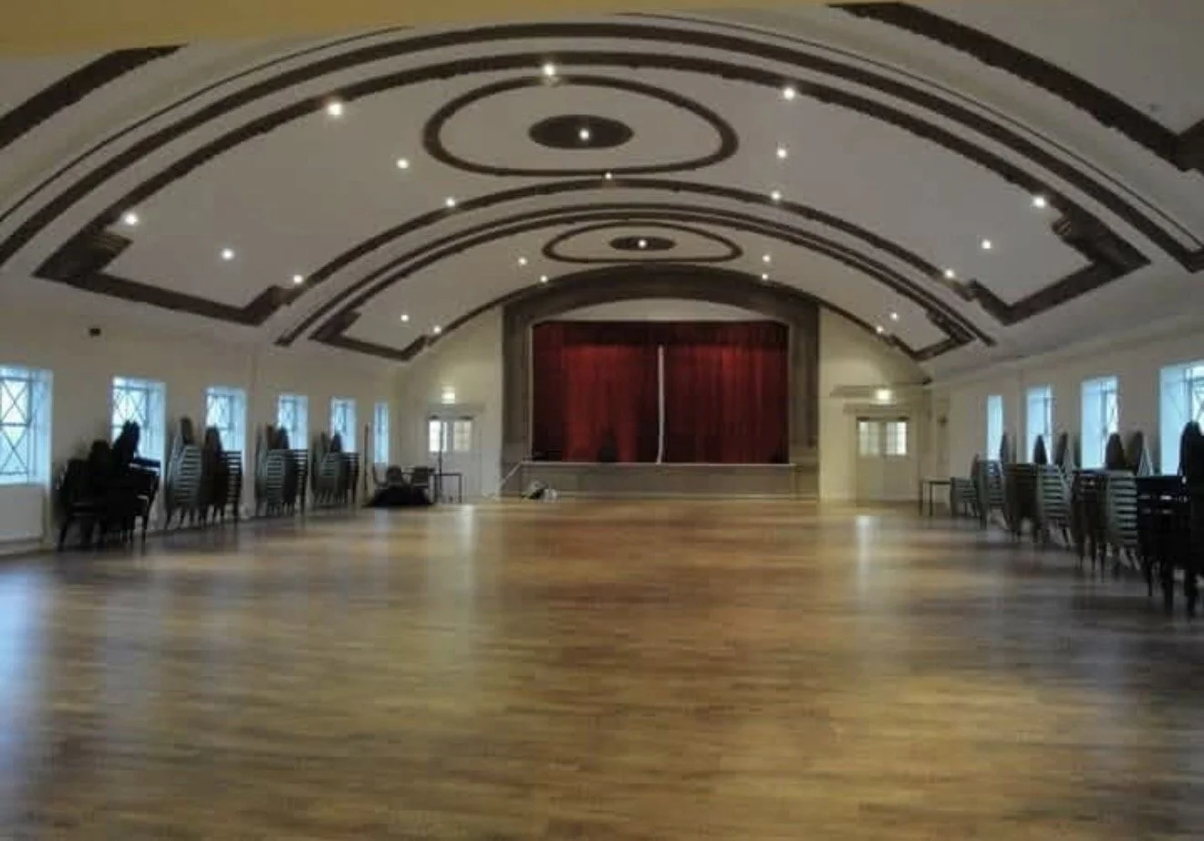 Empty banquet hall or event space with a wooden dance floor, high ceiling with decorative circular patterns, small windows along the walls, and a stage with red curtains at the far end.