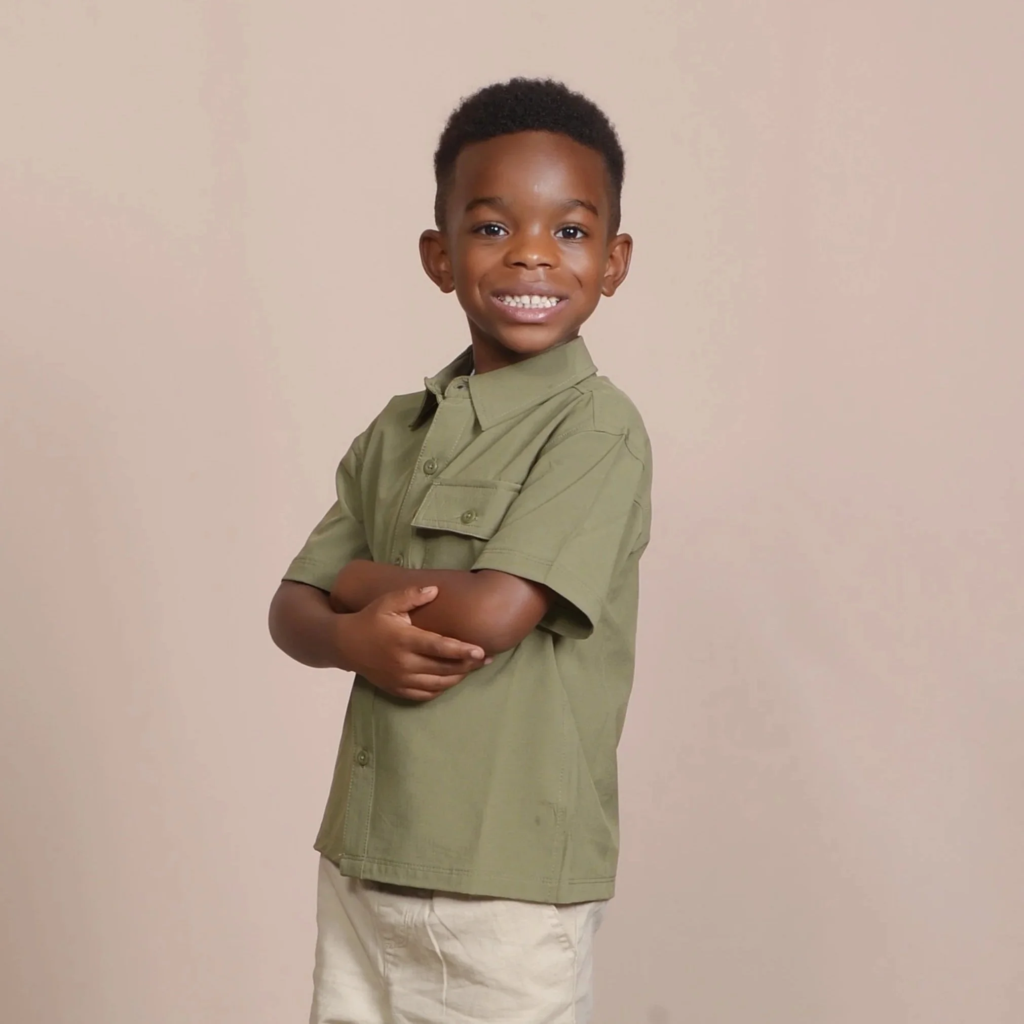 A young boy with short curly hair smiling and crossing his arms against a plain beige background, wearing a light olive green shirt and beige pants.