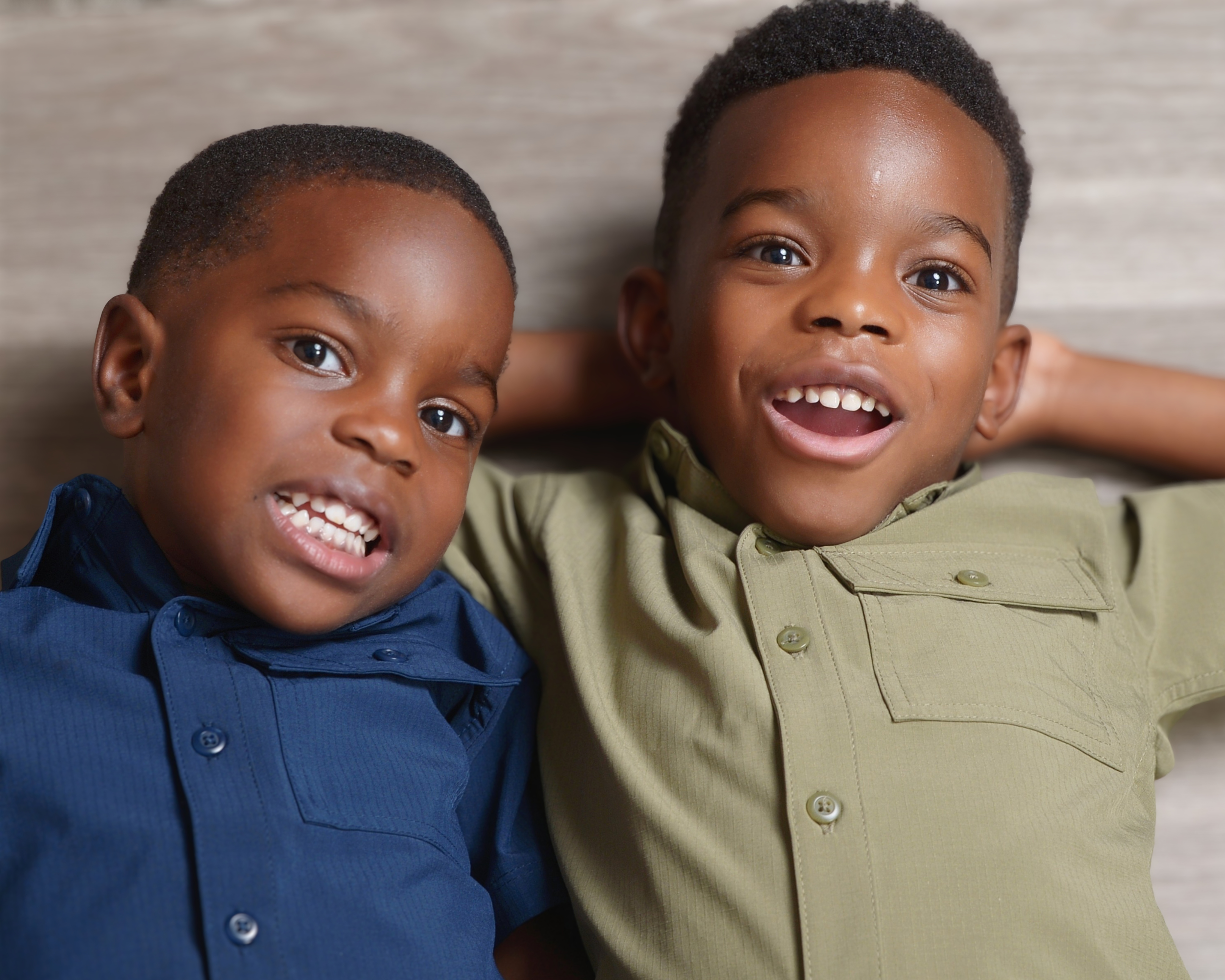 Two young boys with short hair smiling and lying on a light-colored wooden floor, wearing colorful shirts, with one boy wearing a dark blue shirt and the other a light olive green shirt.