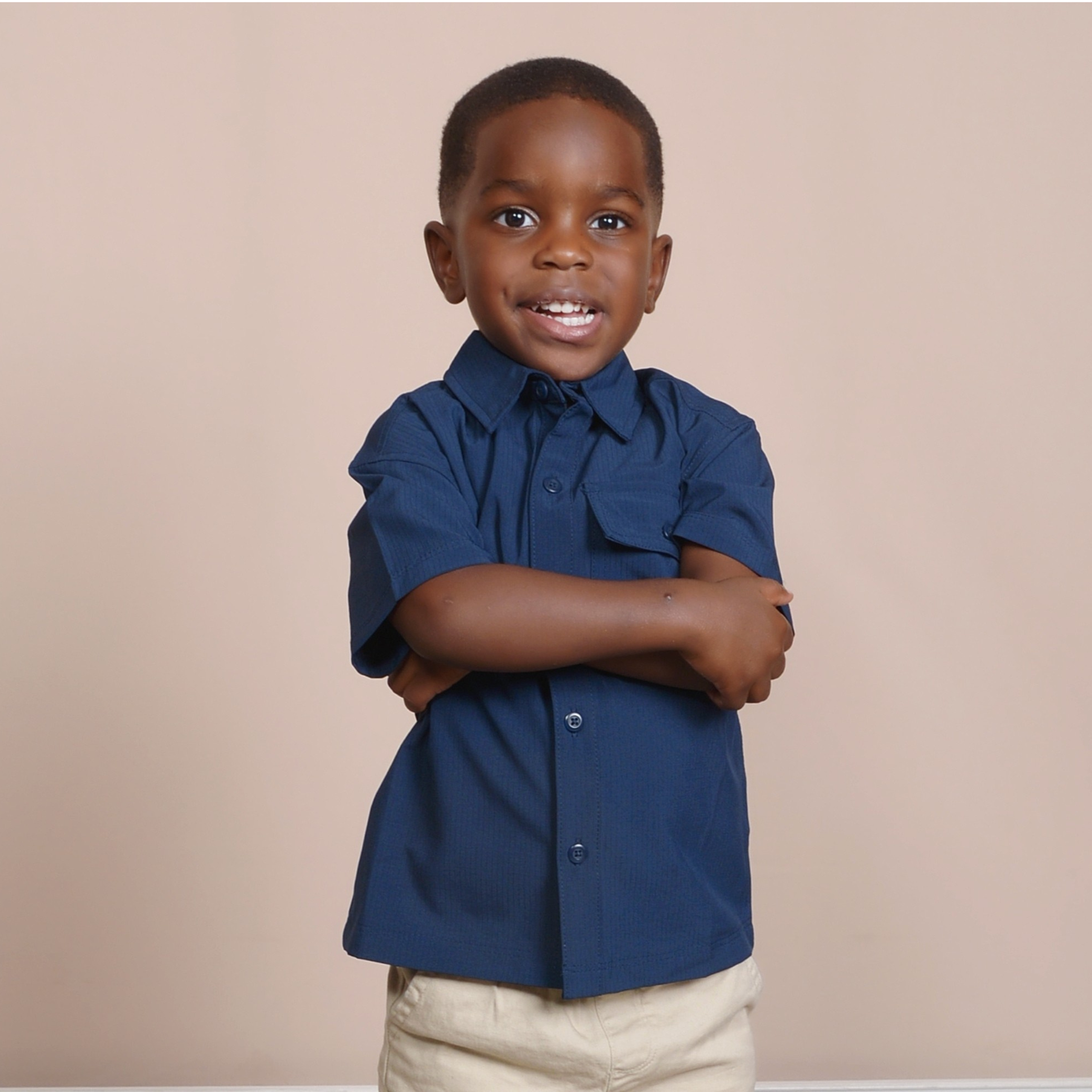 A young boy wearing a dark blue button-up shirt and beige khakis standing against a plain beige wall. He has short black hair and is smiling slightly with his arms crossed.