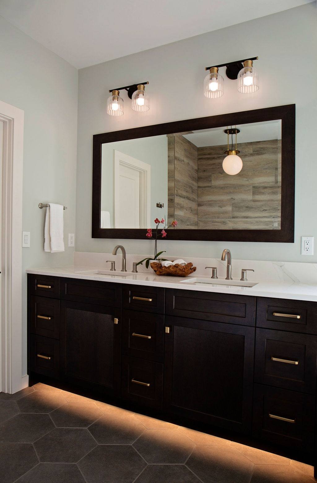 Onyx stained custom Dura Supreme vanity with with reeded doors and toe kick lighting, mixed metal sconces and large custom  framed mirror to match vanity finish reflecting wood-look tile tub accent wall, Pleasant Hill MO