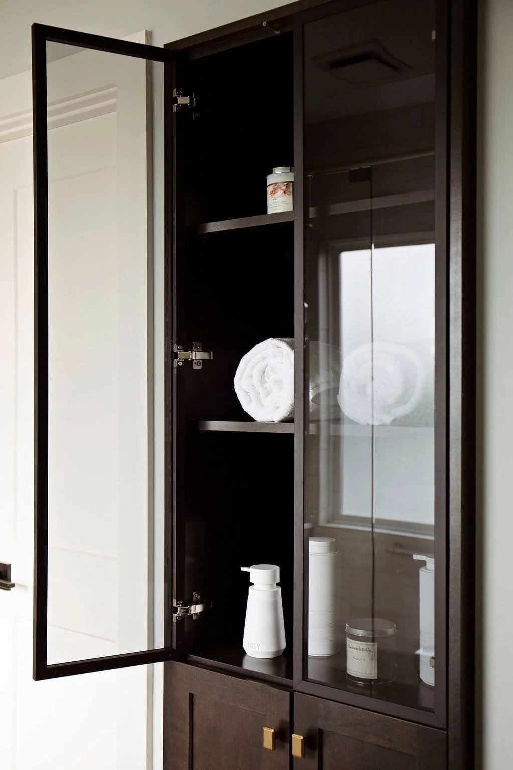 Glass-front storage cabinet with aluminum framed doors with folded towels in onyx cherry wood linen closet, Pleasant Hill MO primary bath renovation. 