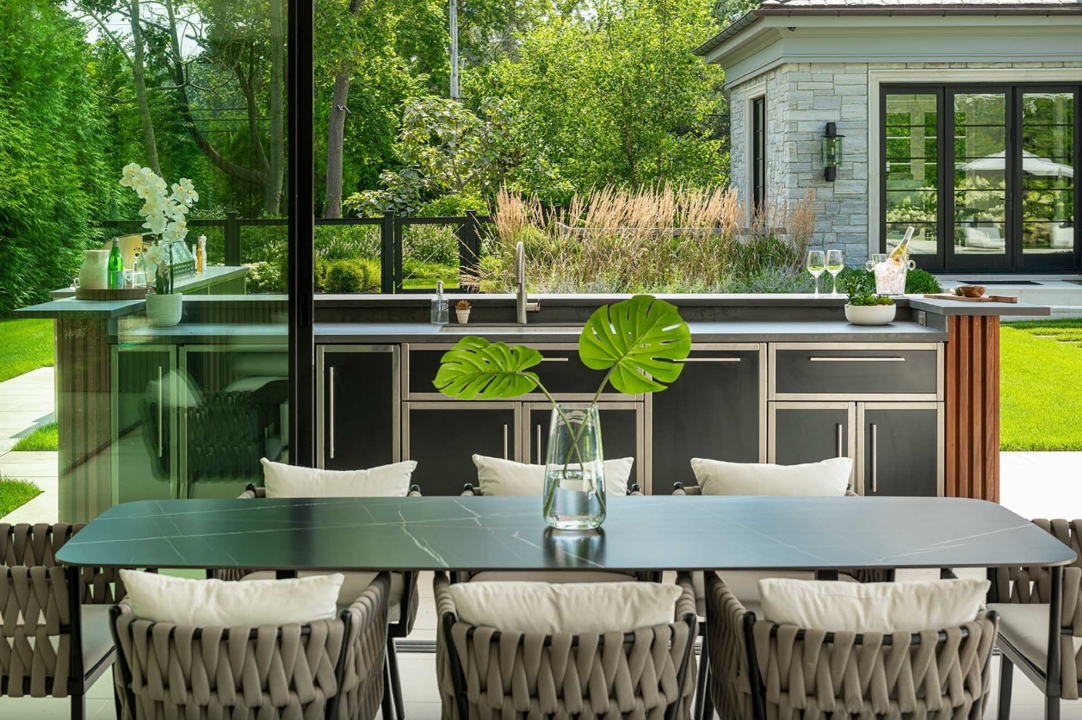 Exterior view of an outdoor kitchen with marine-grade stainless steel cabinetry, outdoor grade countertops and dining area with a table and chairs, and a lush green backyard.