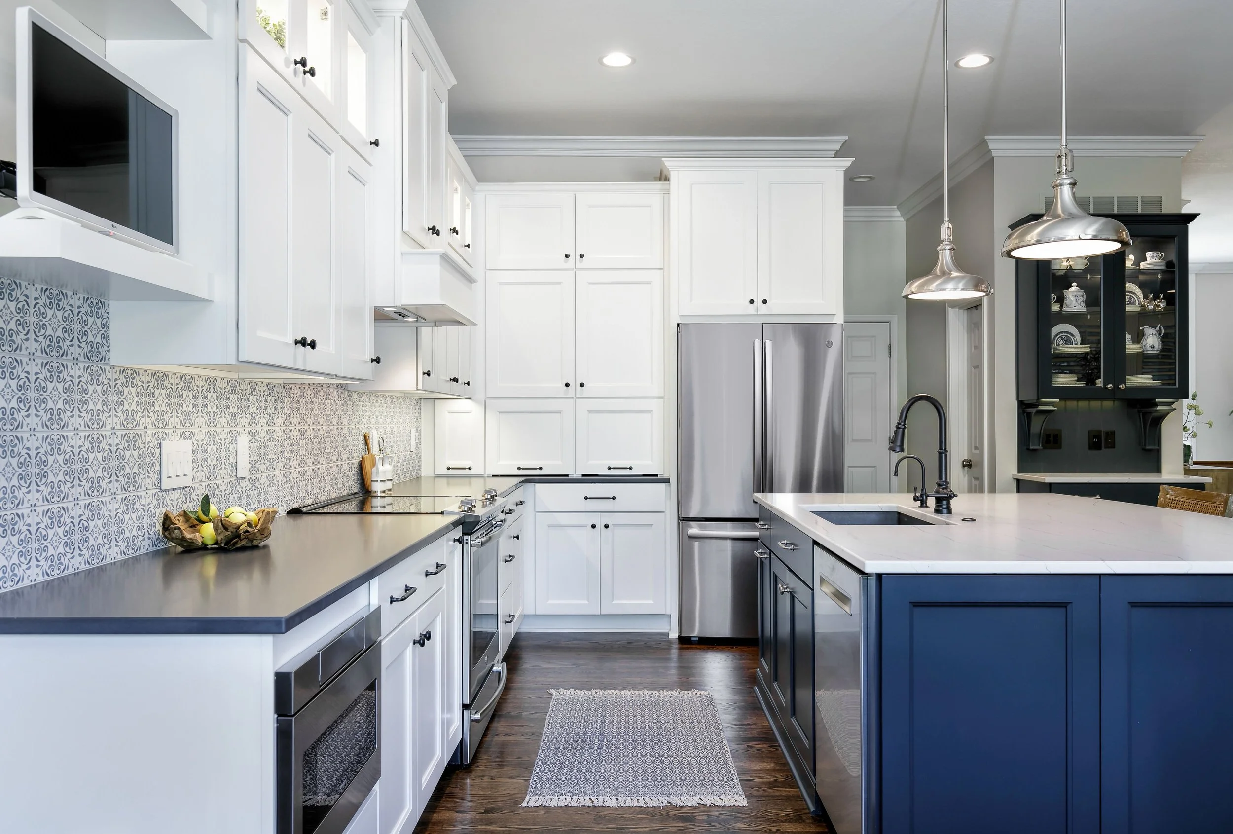 A modern kitchen with white cabinets, black knobs, stainless steel appliances, a patterned backsplash, a wooden bowl with lemons, and a white island with a black faucet. There are pendant lights hanging above the island and a dark wood hutch displayi