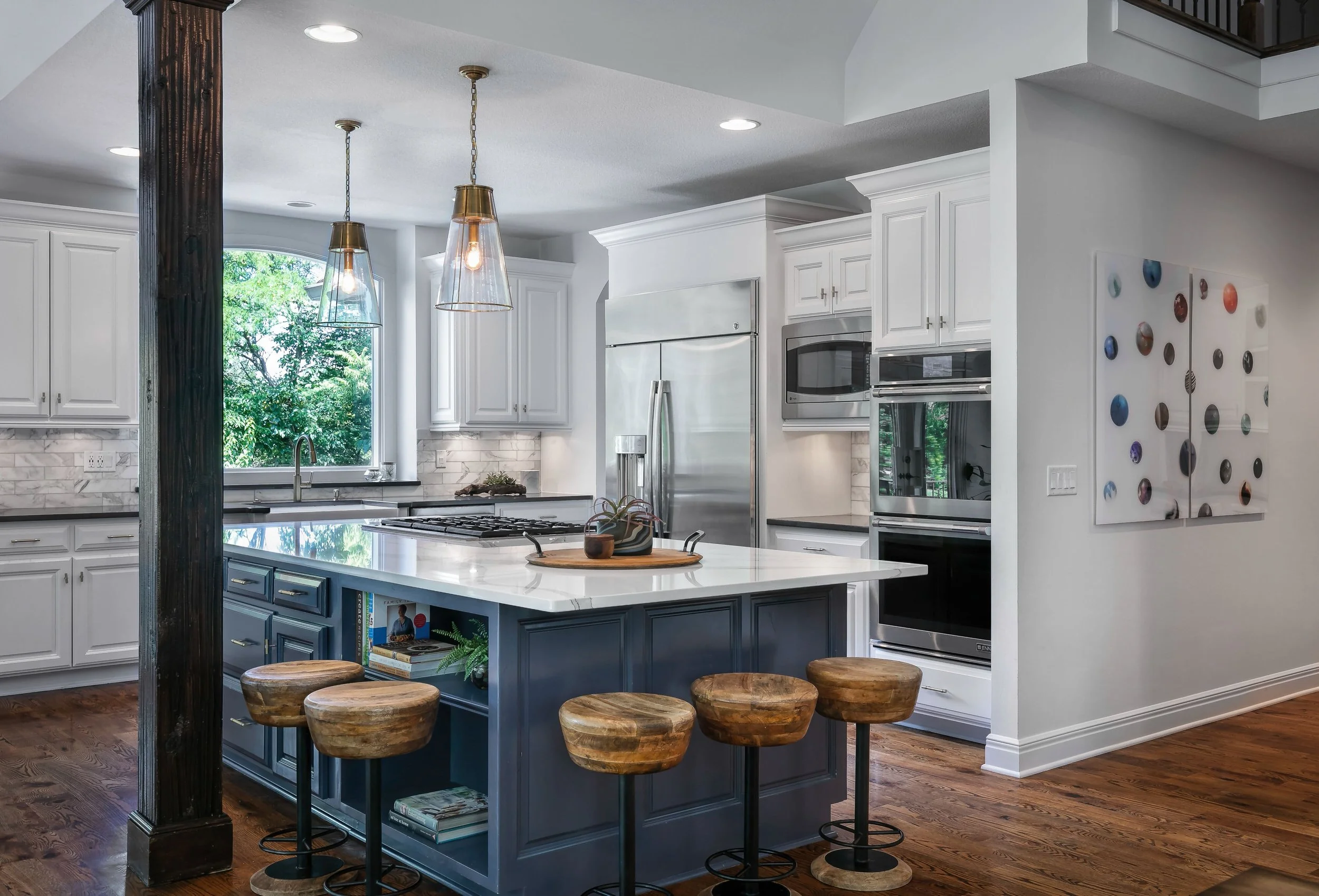 Modern kitchen with white cabinets, stainless steel appliances, and a blue island with books underneath. Wooden stools are arranged around the island, and pendant lights hang above. A window shows greenery outside.