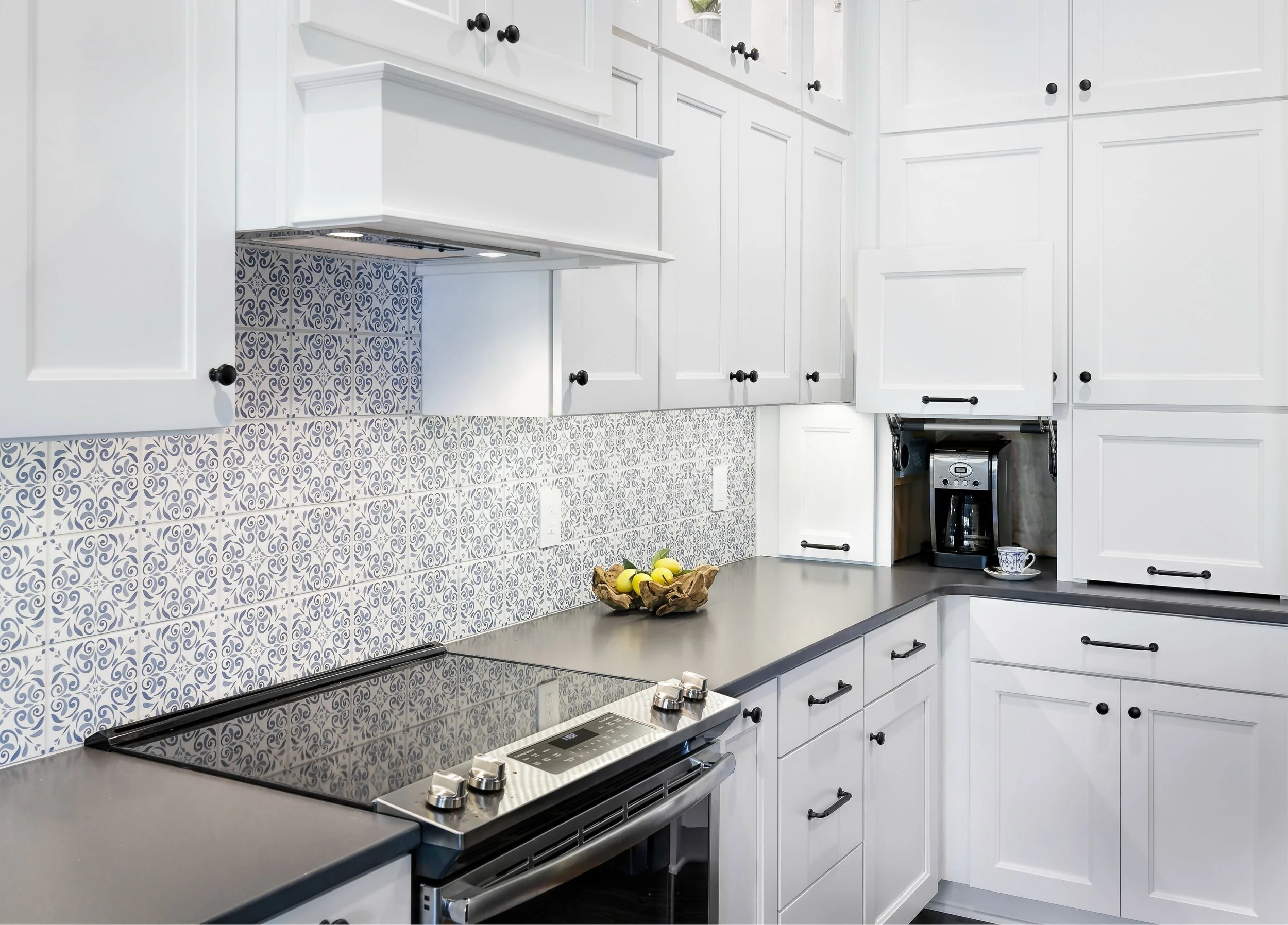 White kitchen cabinets with black knobs, patterned tile backsplash, black stove with digital controls, and a coffee maker on the counter with a mug.