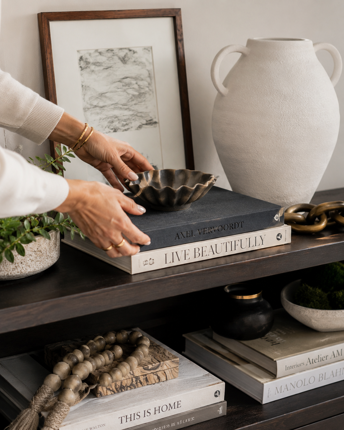 Person arranging decorative items on a dark wooden shelf, including a large white textured vase, a framed black and white art piece, a black ceramic bowl, and a chain, with stacked books underneath.
