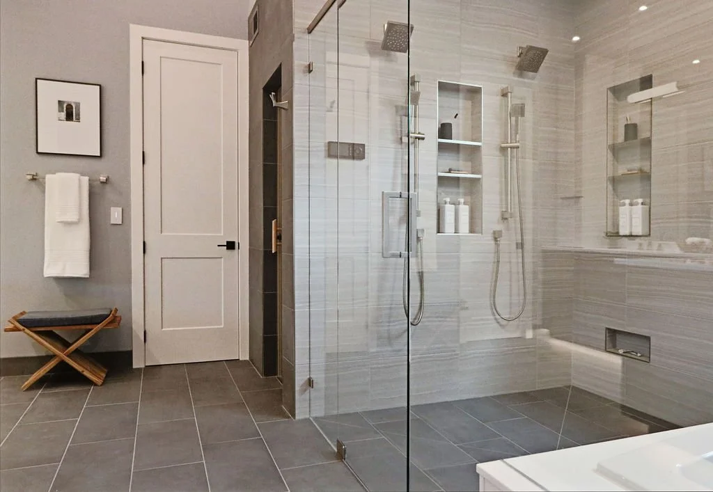 Modern bathroom with a glass-enclosed shower featuring two showerheads, built-in shelves with toiletries, a white door, gray tiled floor, towel rack, and minimal decor.