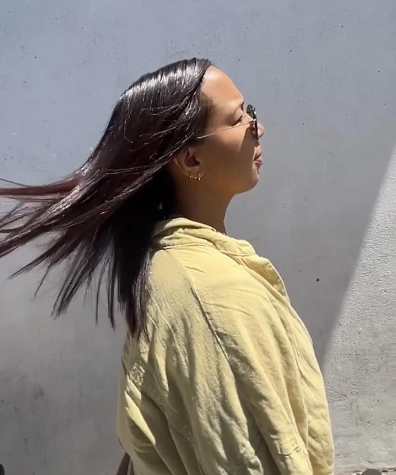A woman with long, dark hair wearing a relaxer and a yellow jacket stands against a grey wall. Her hair is blowing in the wind as she faces to the right.