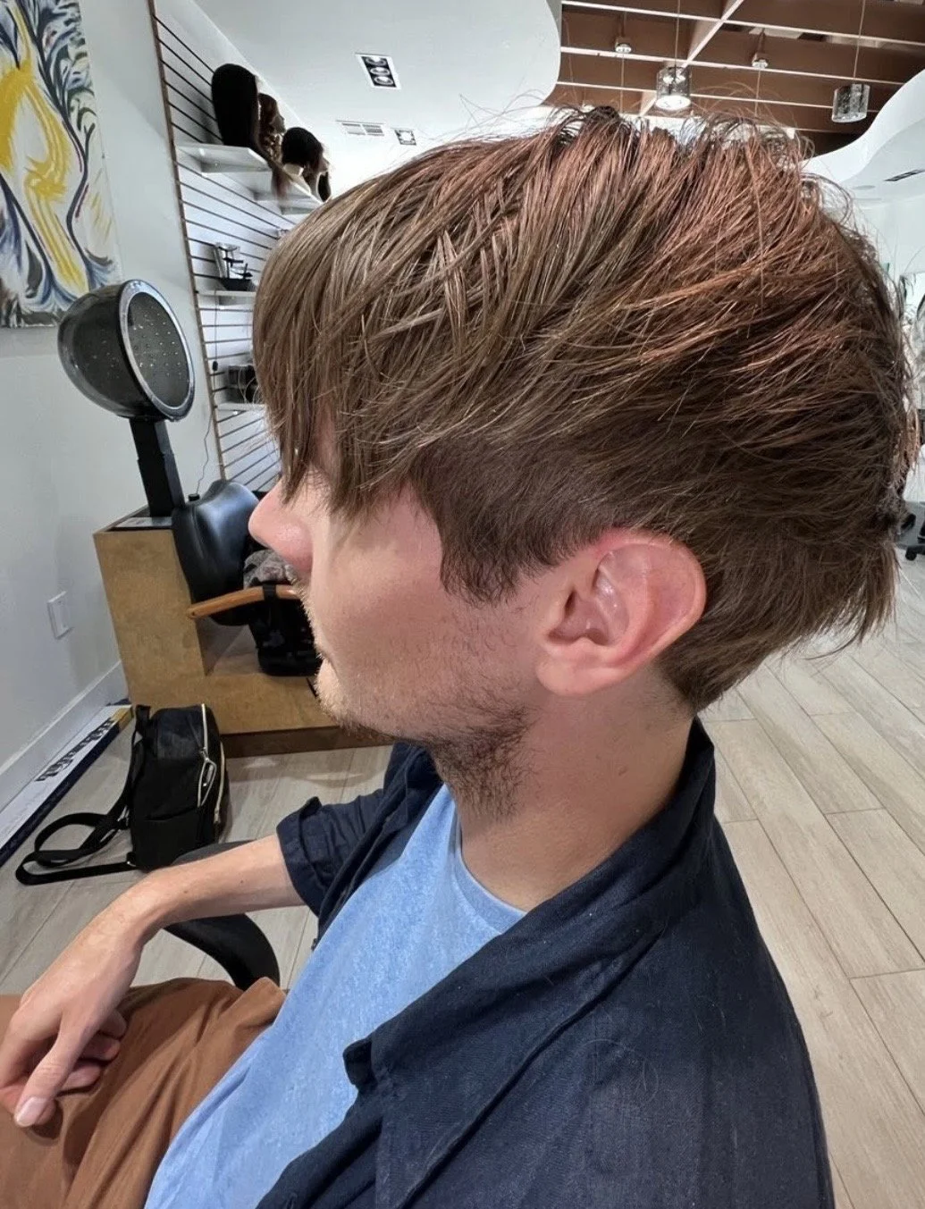 A man getting a haircut at a salon, with styled brown hair and a side profile visible, surrounded by salon equipment and hair products.