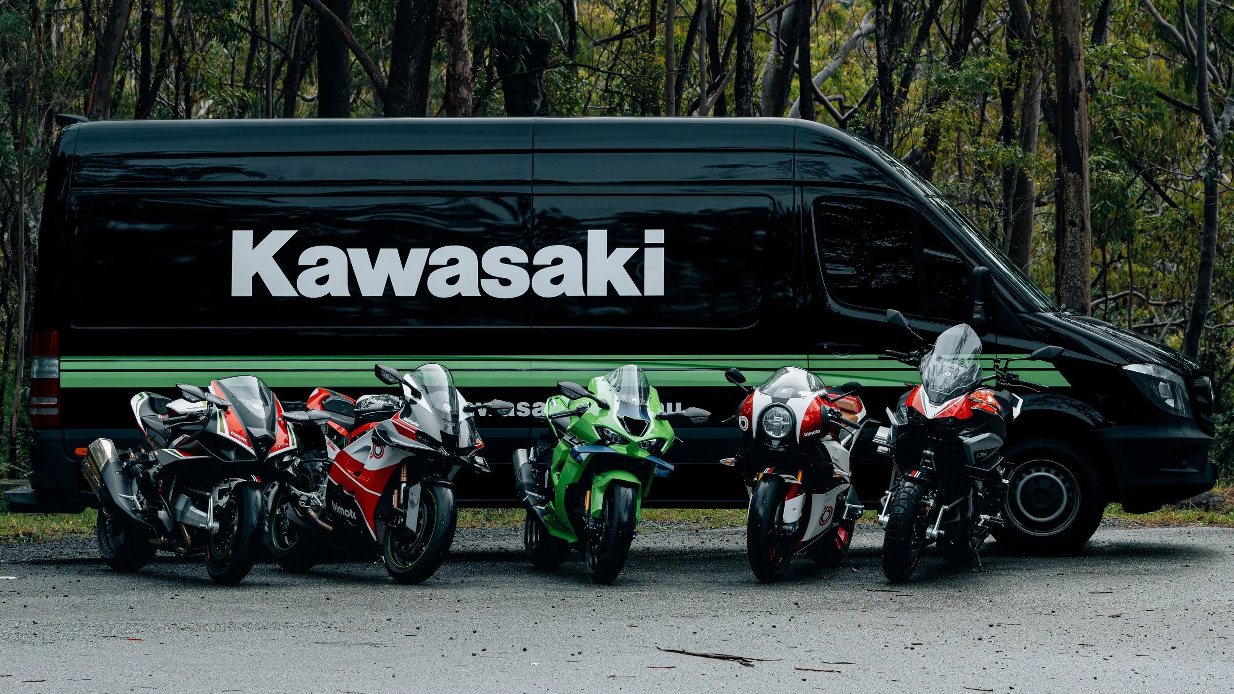A black Kawasaki van parked among several sport motorcycles of various colors, including red, white, green, and black, in a wooded area.