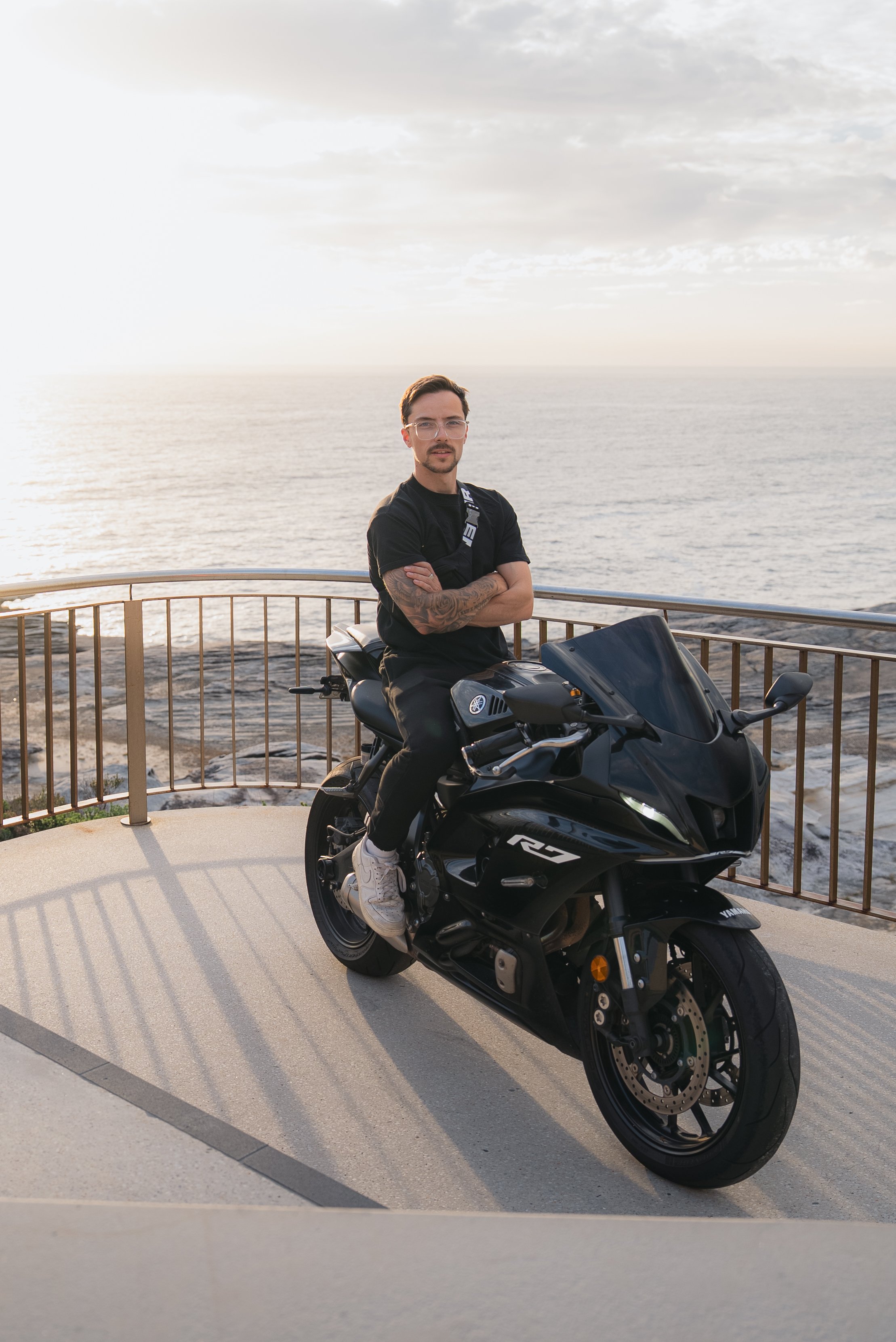 A man with glasses and tattoos sitting on a black Yamaha R15 motorcycle on a lookout point by the ocean during sunset.