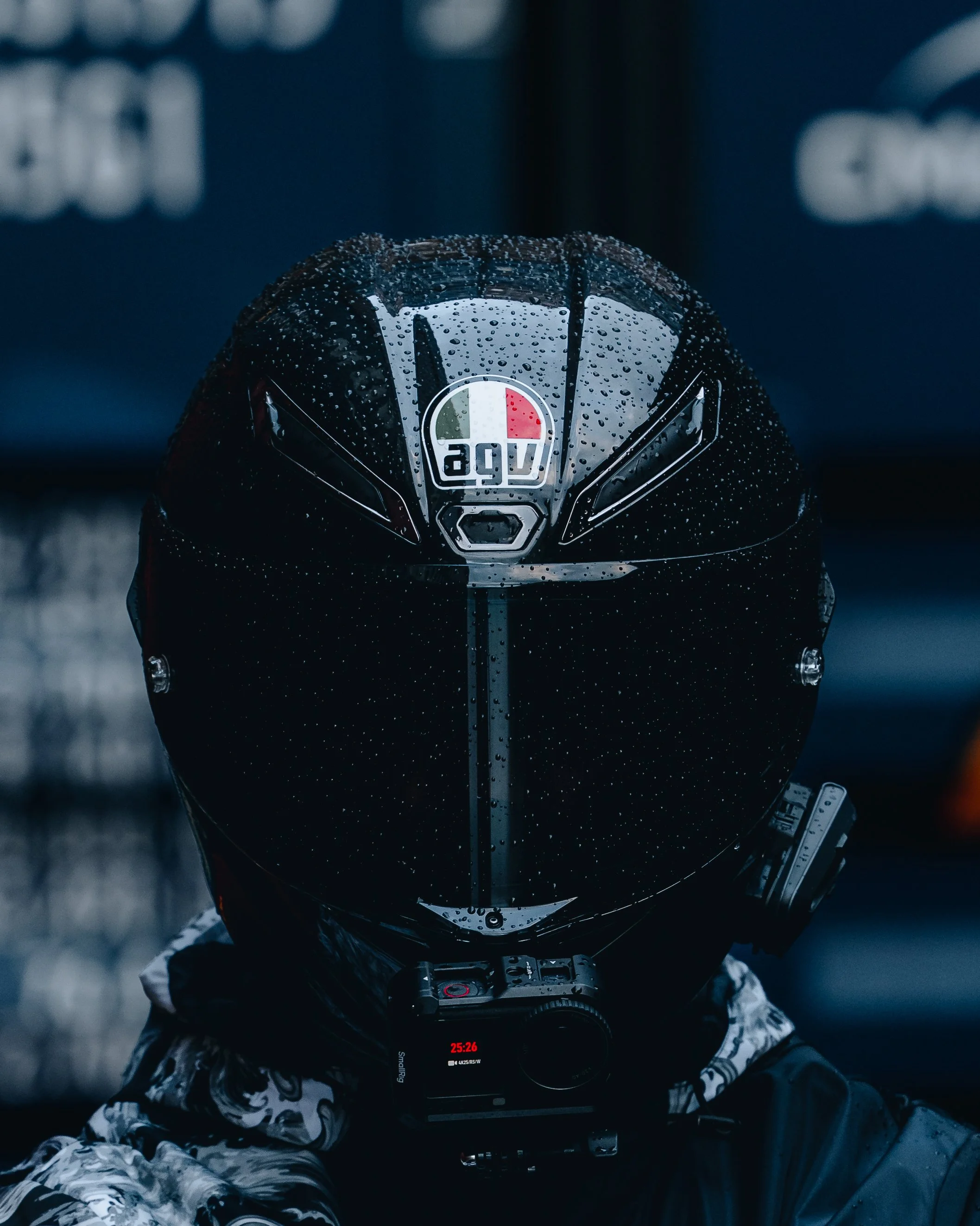 View from above of a person taking a selfie, wearing a black helmet with an 'AGV' logo, and holding a camera or phone, with a rain dropped helmet. The person is dressed in patterned clothing, and the background is blurred.