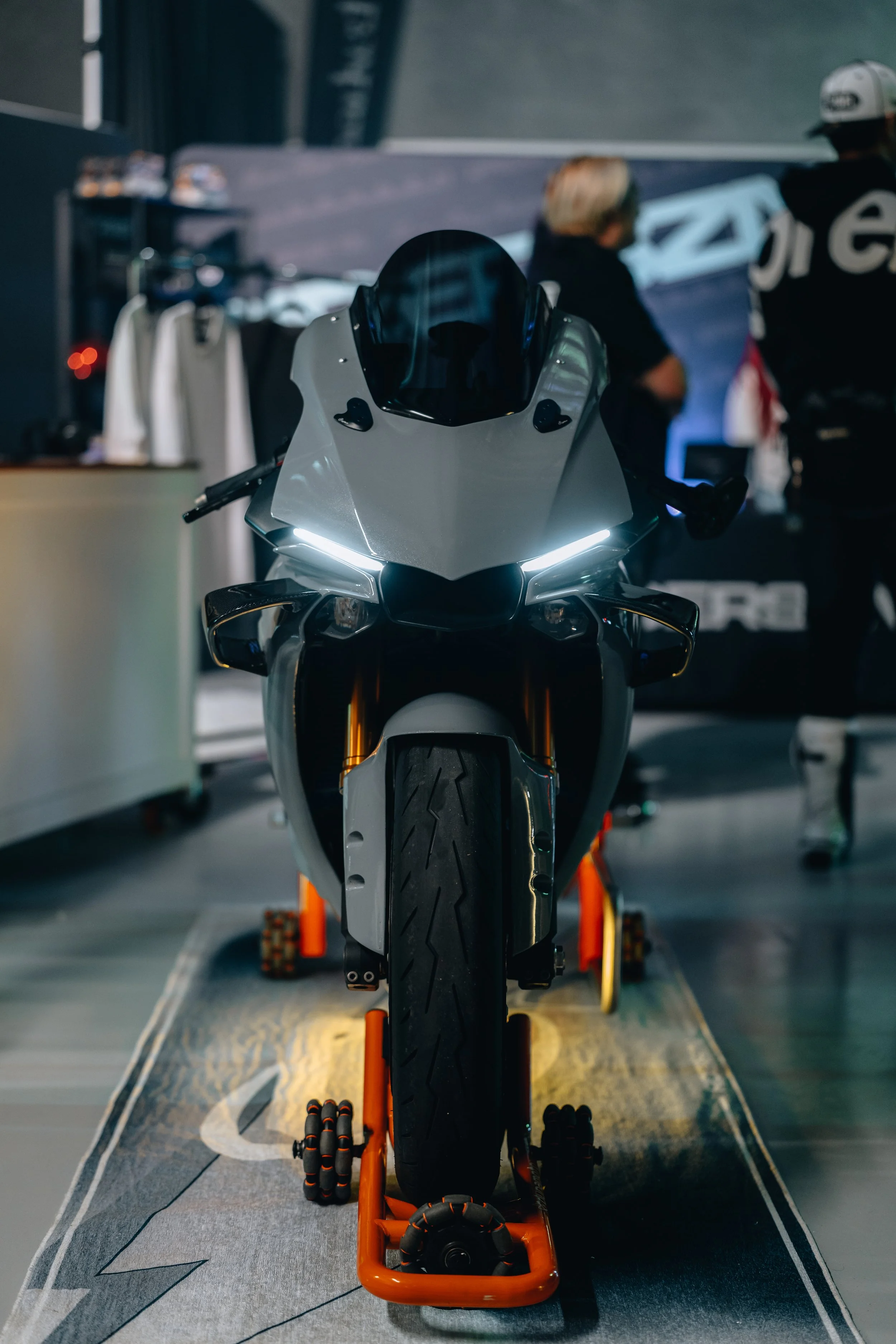A motorcycle on a stand in a showroom or workshop, with people and shelves in the background, illuminated by focused lighting.