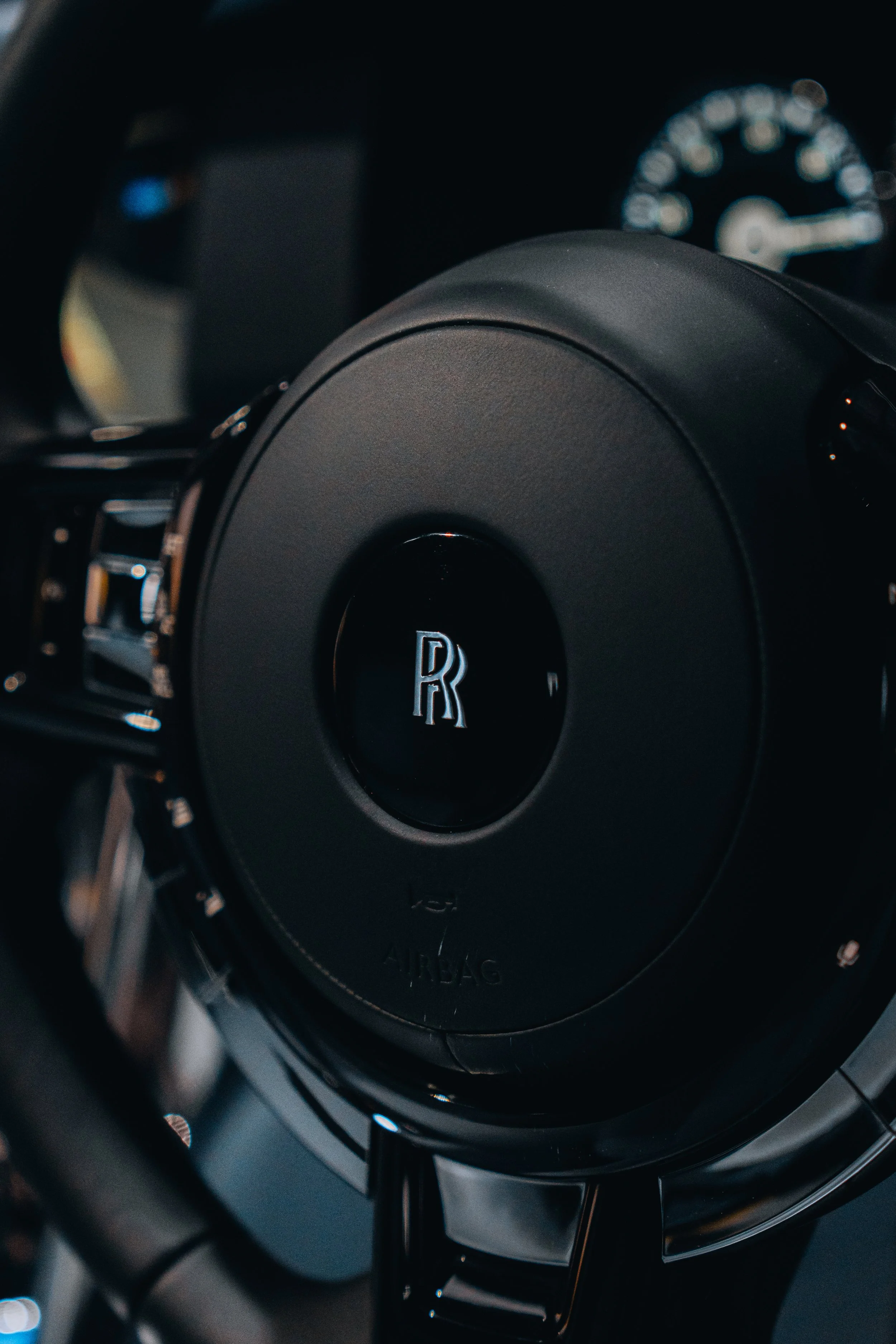 Close-up of the steering wheel of a luxury car, with a Rolls-Royce emblem in the center and a speedometer in the background.