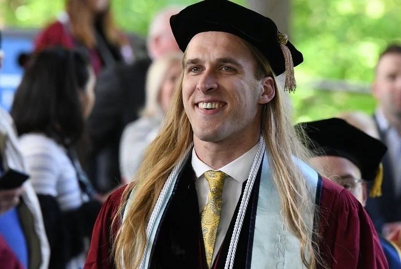 A young man with long blonde hair, wearing graduation regalia including a cap and gown, smiling outdoors at a graduation ceremony.