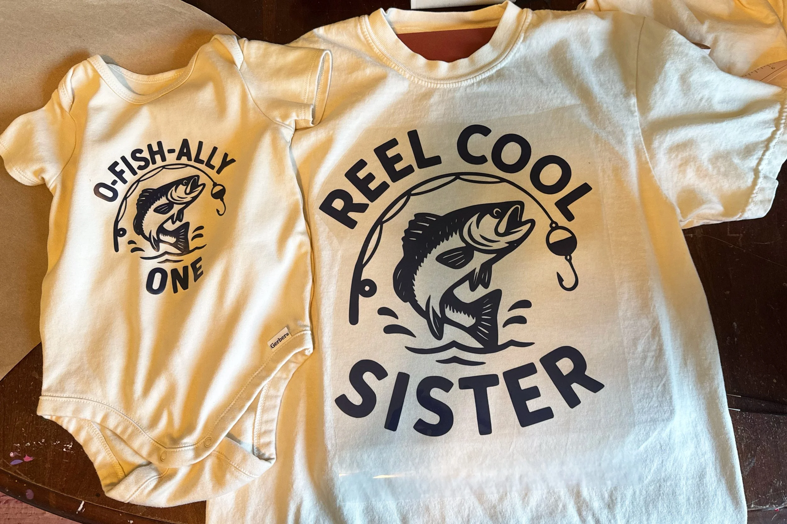 An ivory baby onesie that says "O Fish-ally one" in dark blue with a fish and a fishing pole, and a matching ivory color shirt with dark blue writing saying, "Reel Cool Sister" and matching fish and fishing pole in the same dark blue.