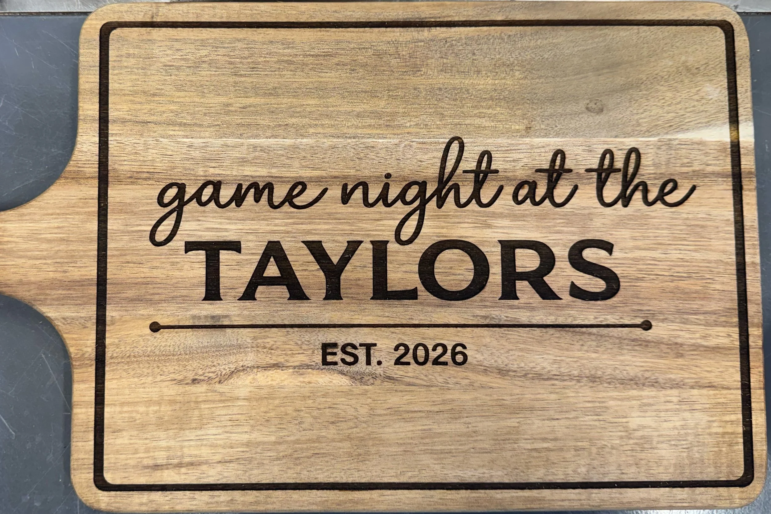 A cutting board engraved with "game night at the Taylors (in all caps) est. 2026"