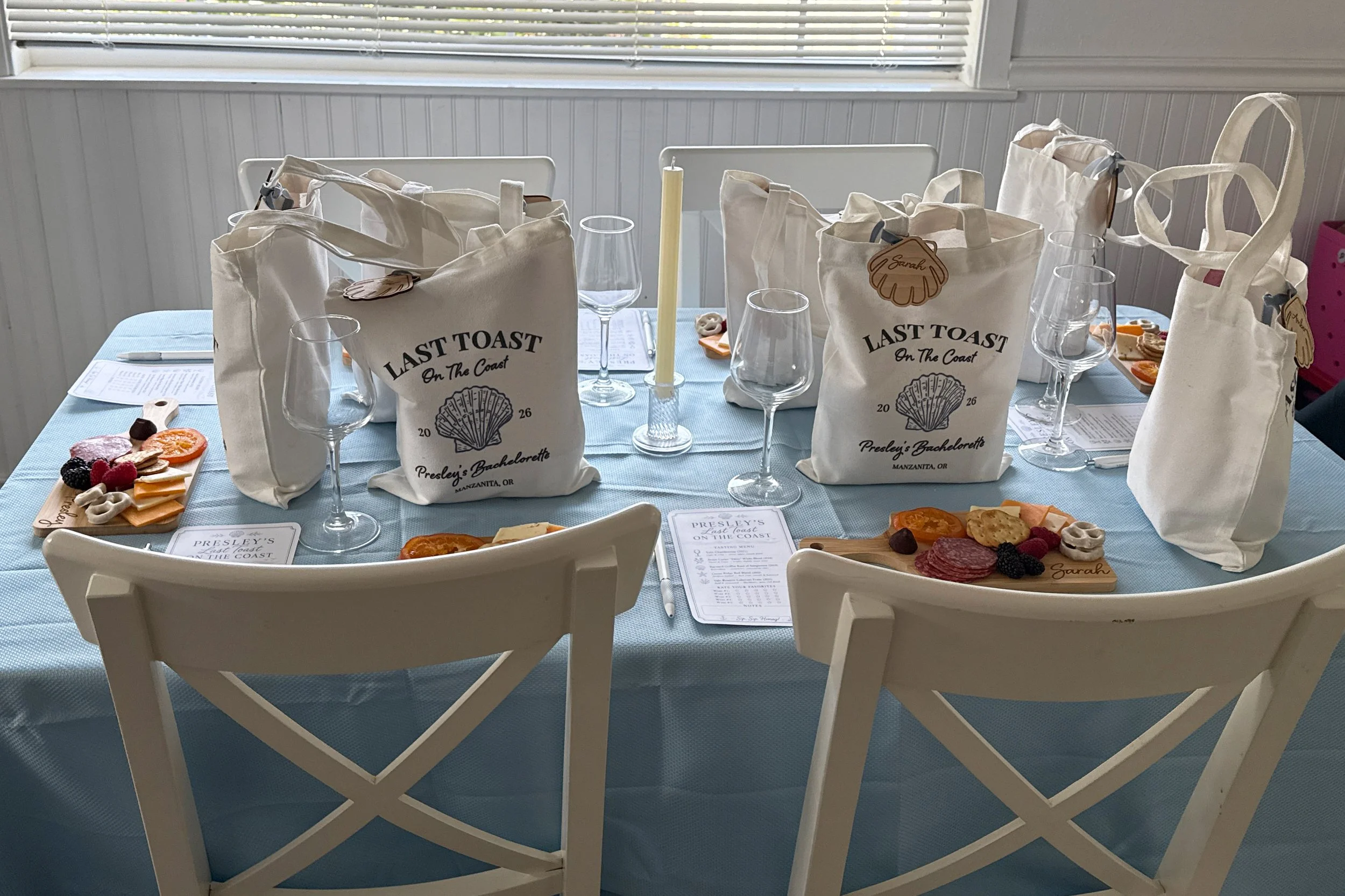 A table set with tote bags saying "Last Toast on the Coast" as well as wine glasses and personal sized charcuterie boards with names engraved on them. 